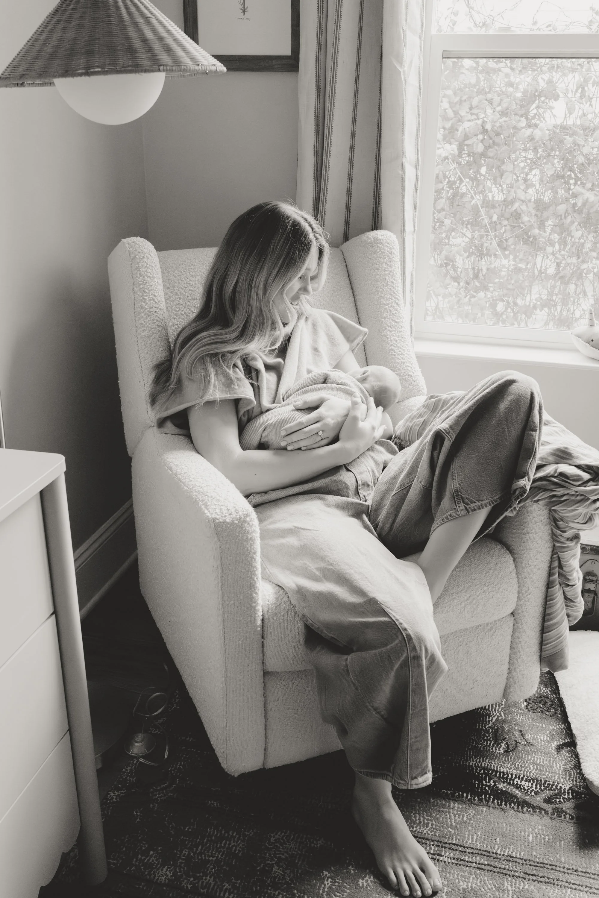 A woman with long wavy hair sitting in a cozy armchair by a window, breastfeeding a baby with care and attention, in a softly lit room. lifestyle newborn photography in Austin 