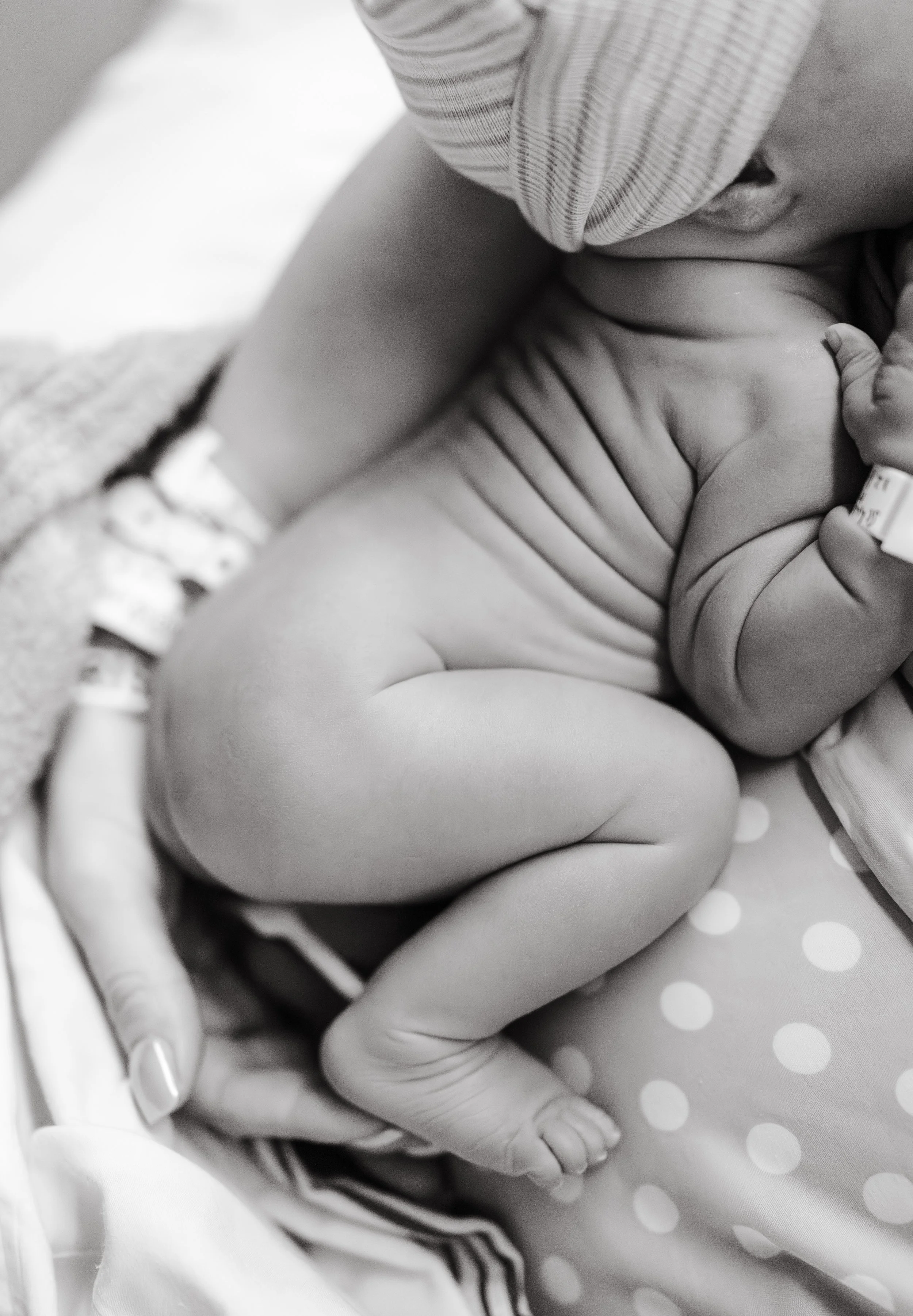A close-up black-and-white photo of a newborn baby lying on a polka dot blanket, with an adult's hand gently holding the baby's tiny foot.