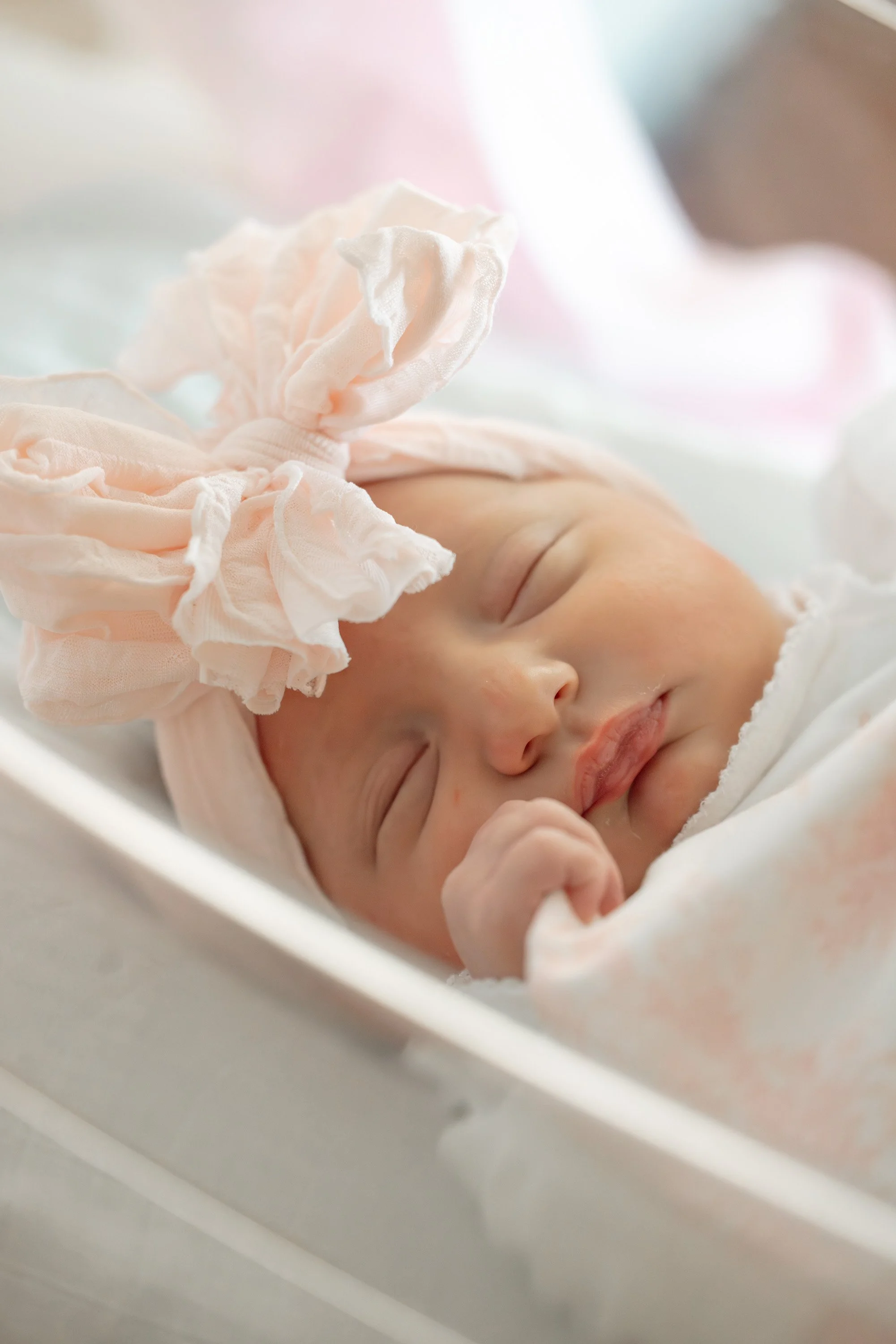 Close-up of a sleeping newborn baby with a soft pink bow around head, resting peacefully in a white crib or bassinet. Austin fresh 48 photography 
