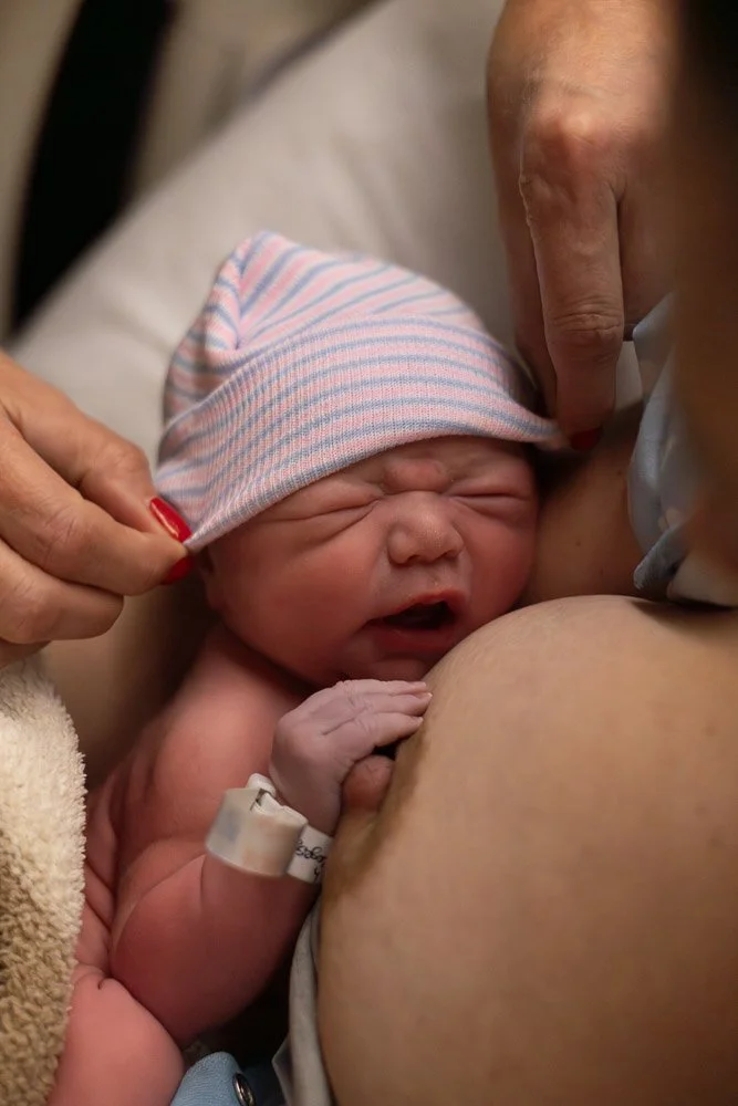 A newborn baby in a hospital setting, wearing a pink and blue striped hat, being held and supported by an adult, likely during or shortly after birth. top rated birth photographer in Austin Texas 