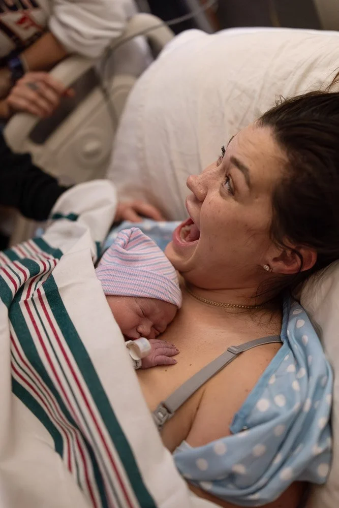 A woman in a hospital bed holding a newborn baby dressed in a pink and blue striped hat, with a hospital blanket nearby. Her facial expression is joyful and surprised.
