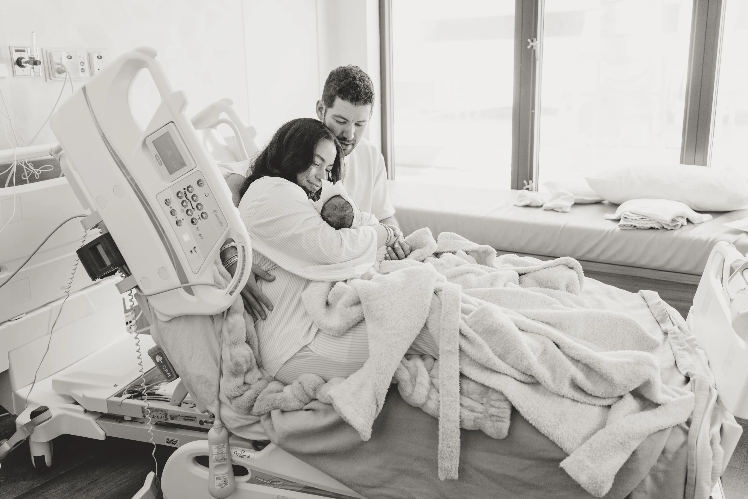 A woman holding a newborn baby in a hospital bed, with a man sitting close beside them, in a hospital room with medical equipment and large windows. fresh 48 photo session Austin Texas