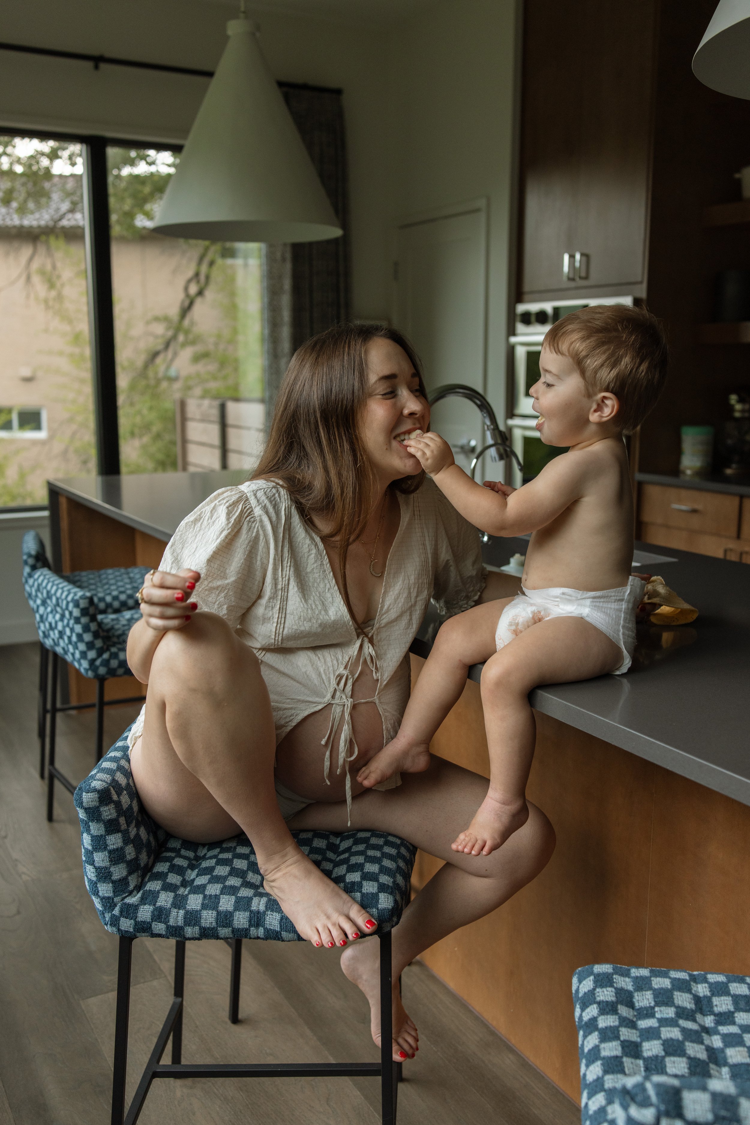 A woman and a toddler sitting on a kitchen counter, playing and smiling; woman sitting on a chair with legs crossed, woman has brown hair, and the toddler has light brown hair, both with light skin. The woman is wearing a beige top, and the toddler i