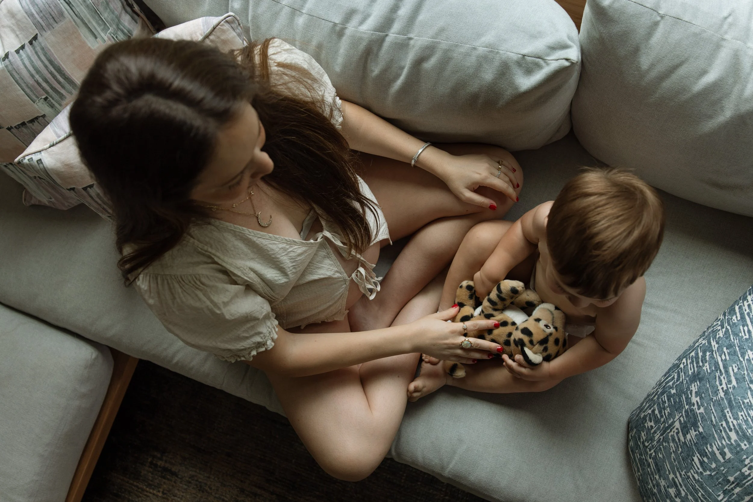 A pregnant woman with long brown hair lying on a gray sofa next to a young boy with light brown hair, holding a stuffed animal tiger. lifestyle maternity session in austin