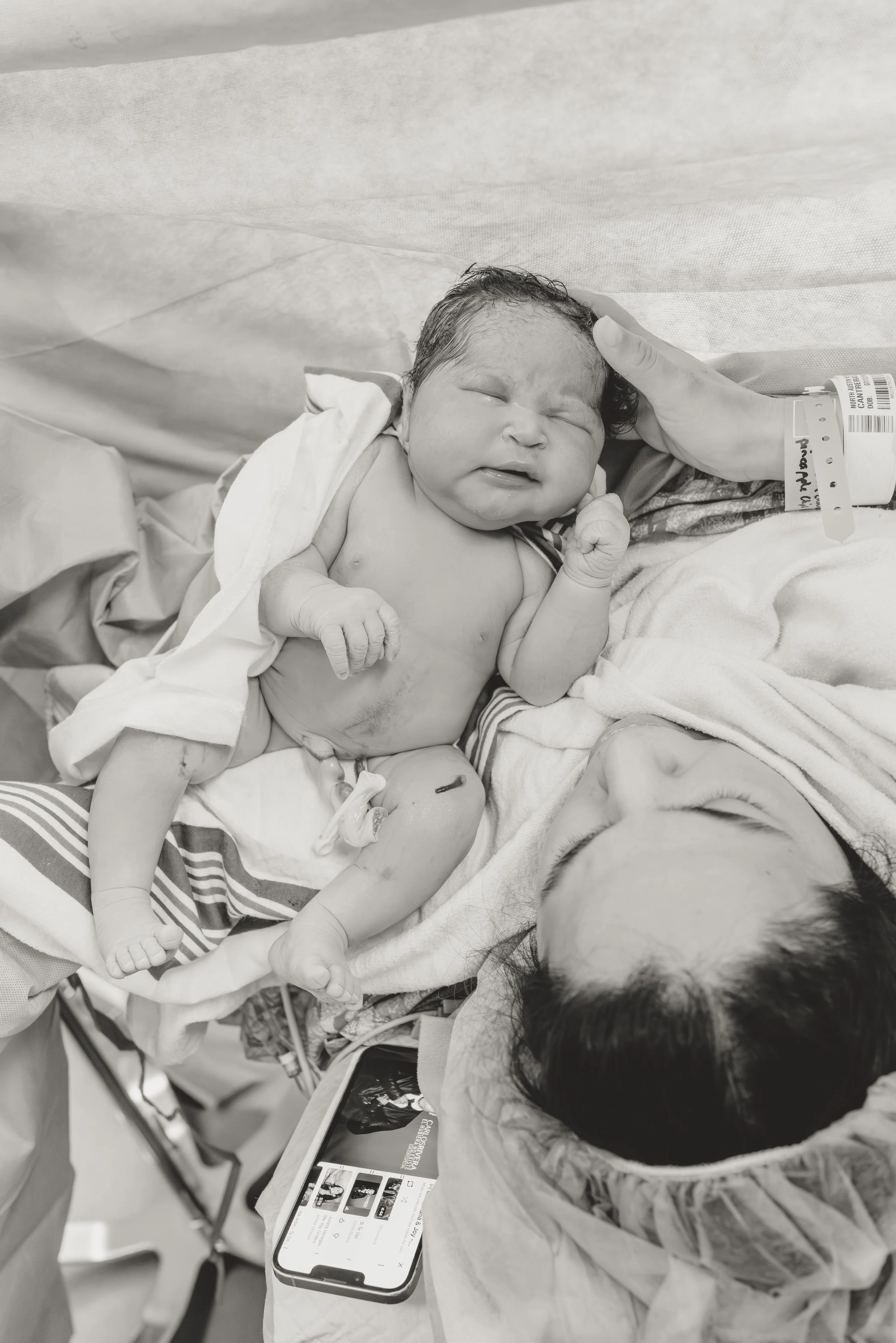 Newborn baby lying on a hospital bed with an adult's hand gently supporting the baby's head, surrounded by hospital equipment and information on a smartphone screen. birth photographer in dripping springs Texas