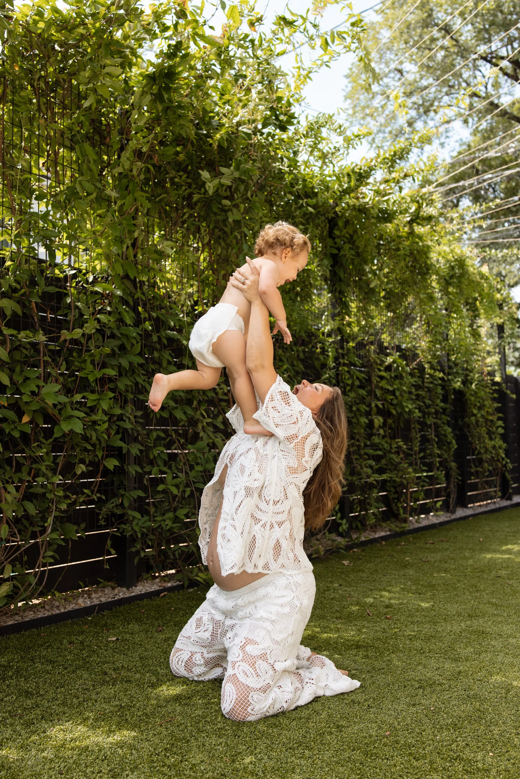 a pregnant woman in a lacy 2 piece set sits outside on grass holding her toddler above her head for maternity photos. 