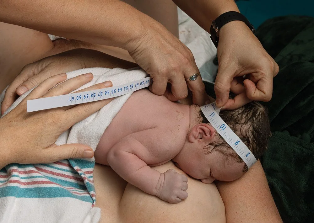 A newborn baby being measured with a measuring tape while lying on a woman's chest, with two hands assisting in the measurement. home birth photographer in Austin Texas