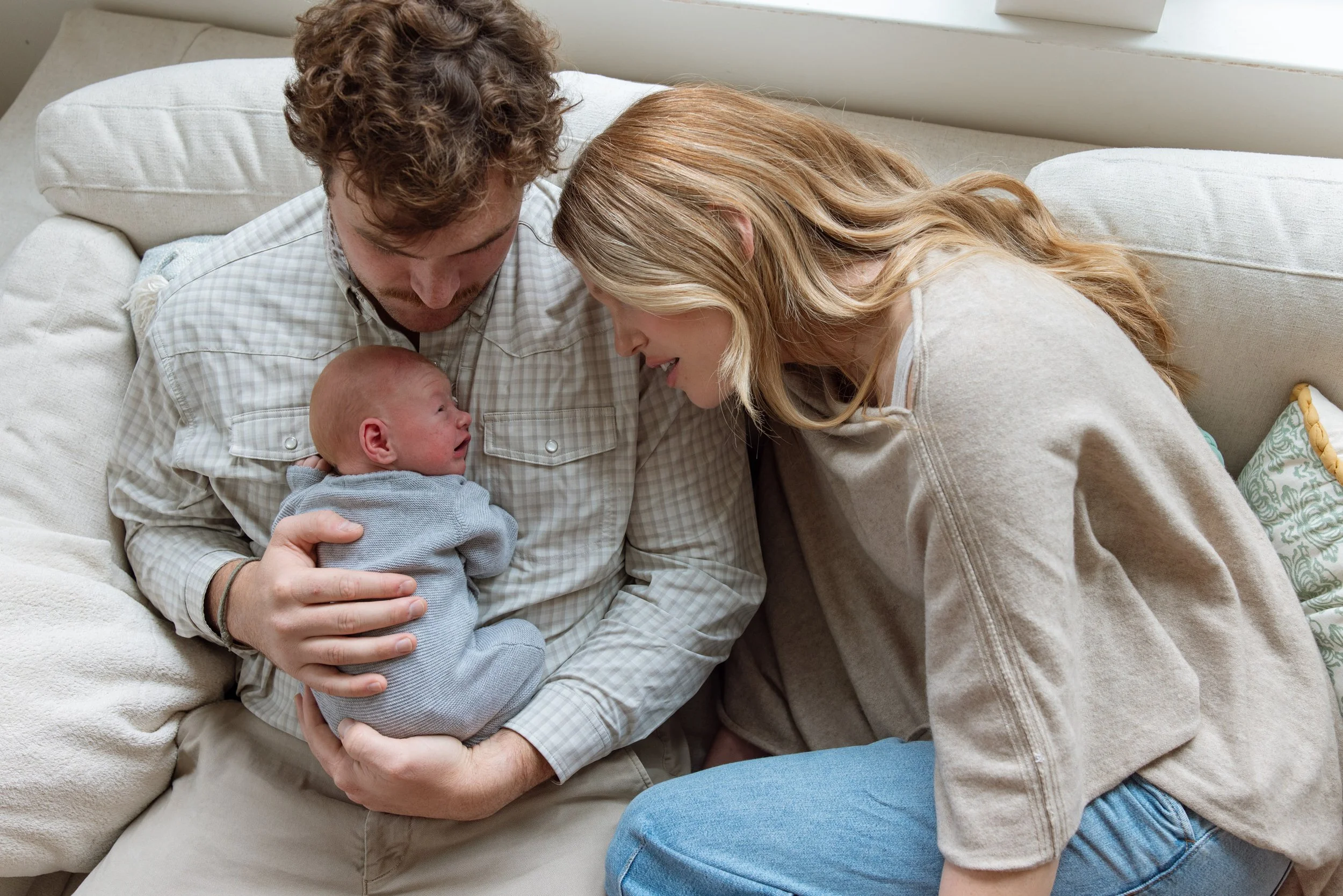 A family of three sitting on a beige couch, with a man holding a newborn baby and the woman leaning in, all looking at each other lovingly.