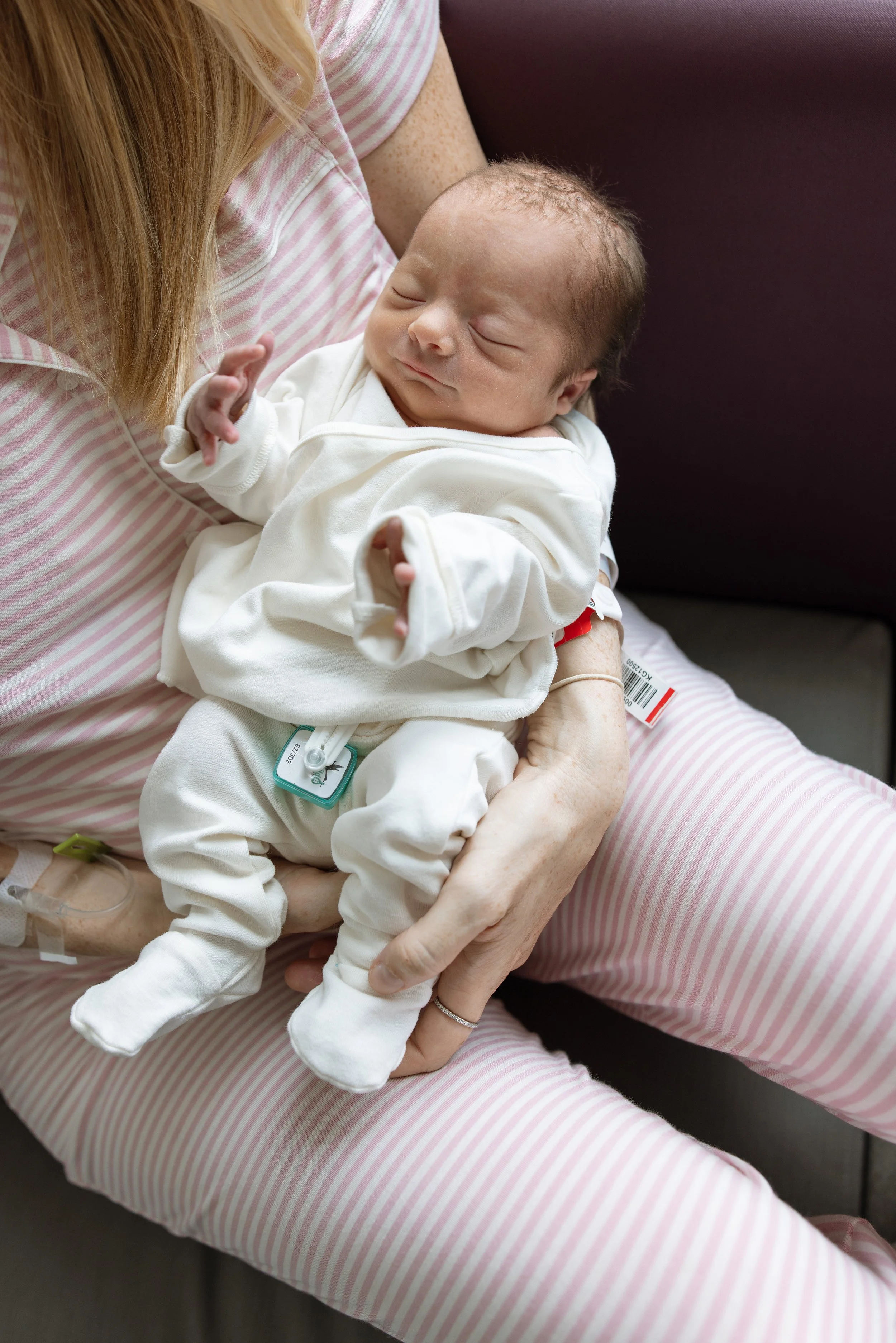 Close-up of a newborn baby sleeping on a woman's lap, who is wearing pink and white striped pajamas, in a hospital setting. fresh 48 photography in hospital in Austin Texas