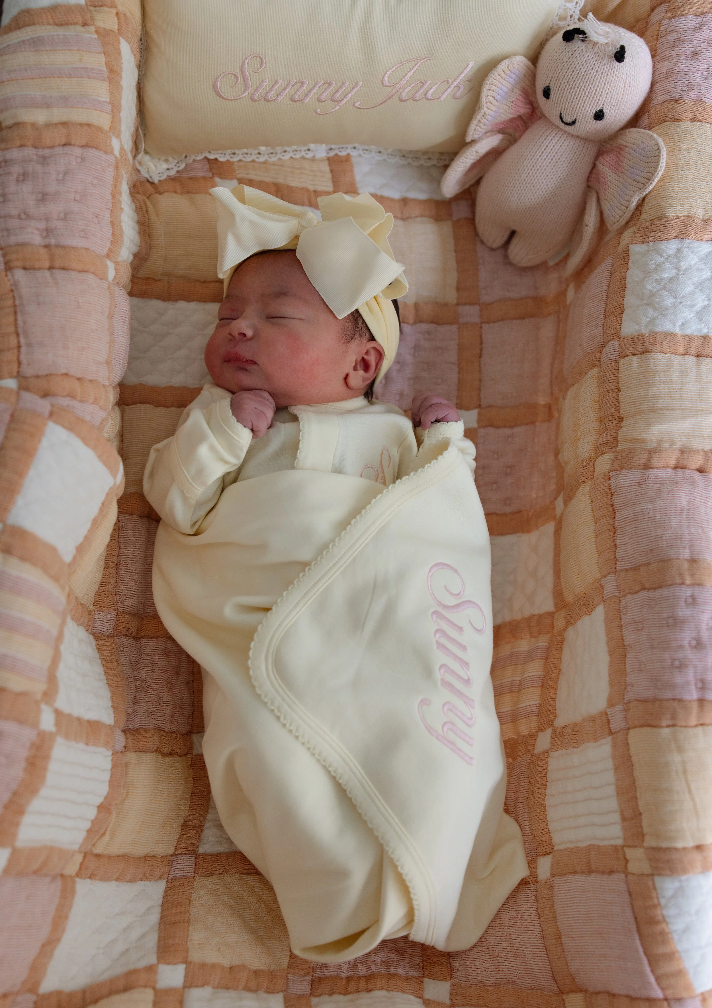 Sleeping newborn baby dressed in cream-colored pajamas and a matching headband with a large bow, lying in a pink and cream quilted bassinet with a pink plush dragon toy nearby and a pillow embroidered with "Sunny Jack." fresh 48 photographer Austin T