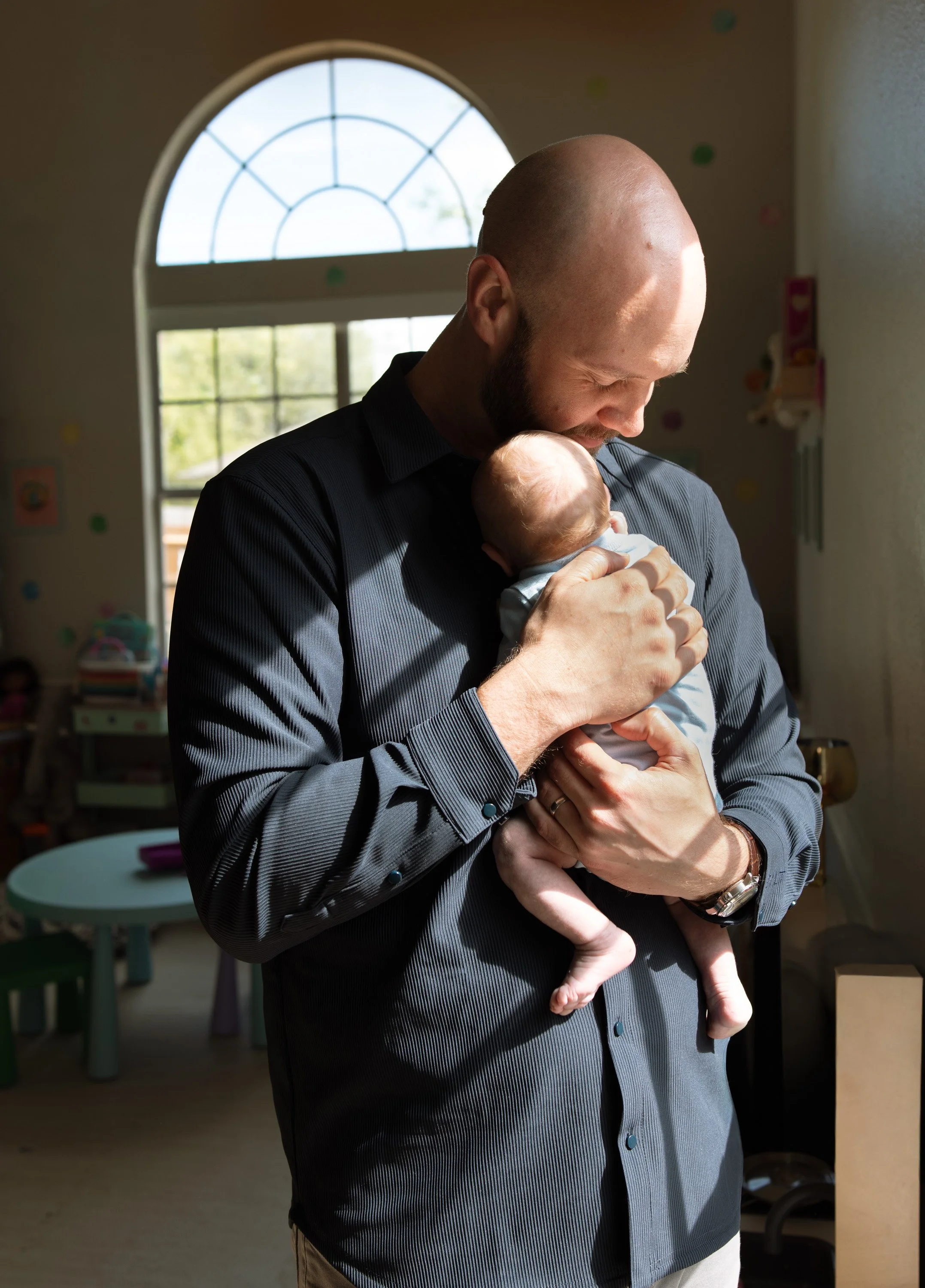 A man with a shaved head and beard holding a baby close to his chest, inside a room with a large window, colorful wall decorations, and child-related furniture. newborn photographer in dripping springs Texas