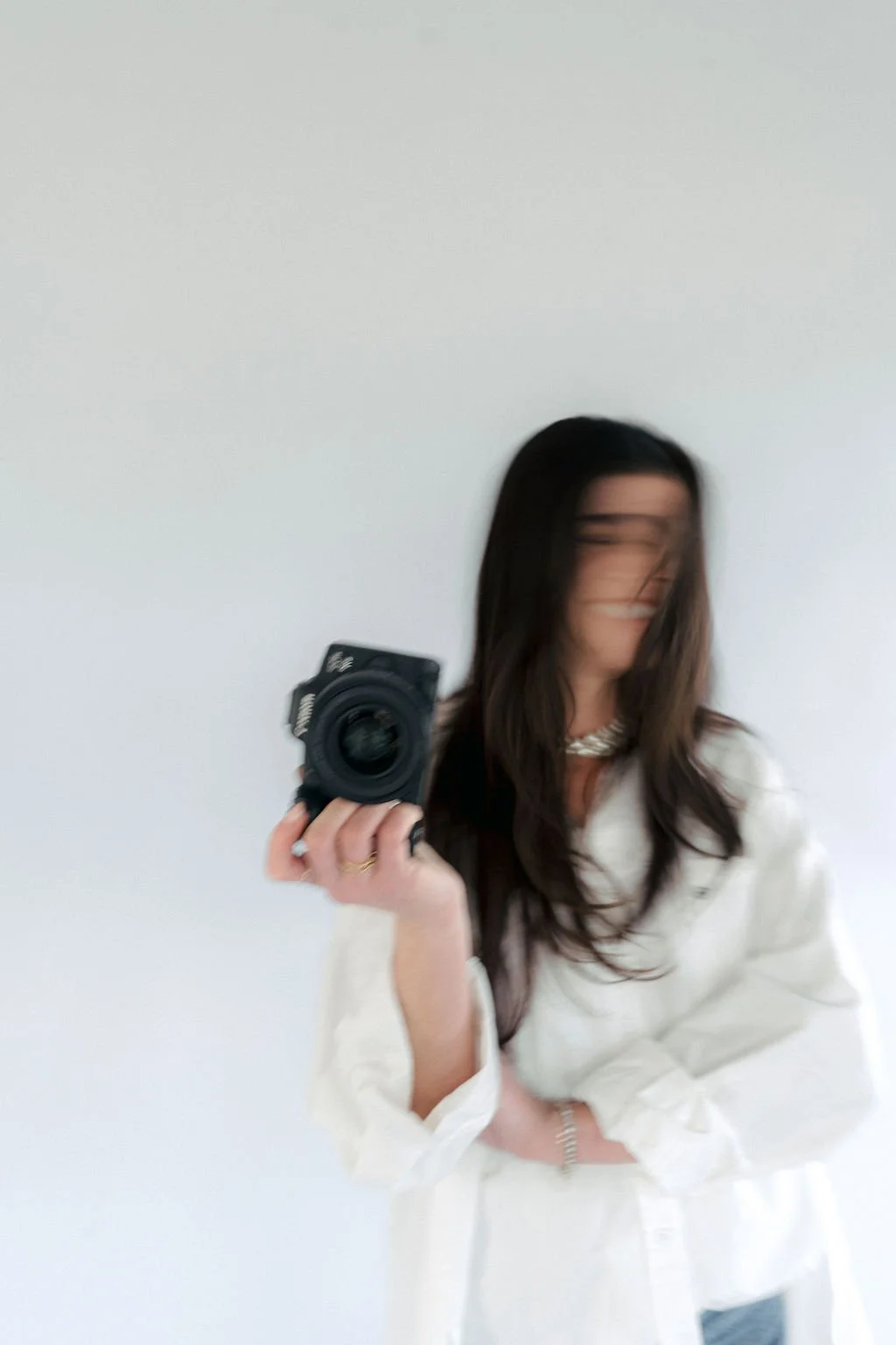 Woman with long brown hair taking a mirror selfie with a camera, wearing a white outfit and a necklace, blurred face and background.