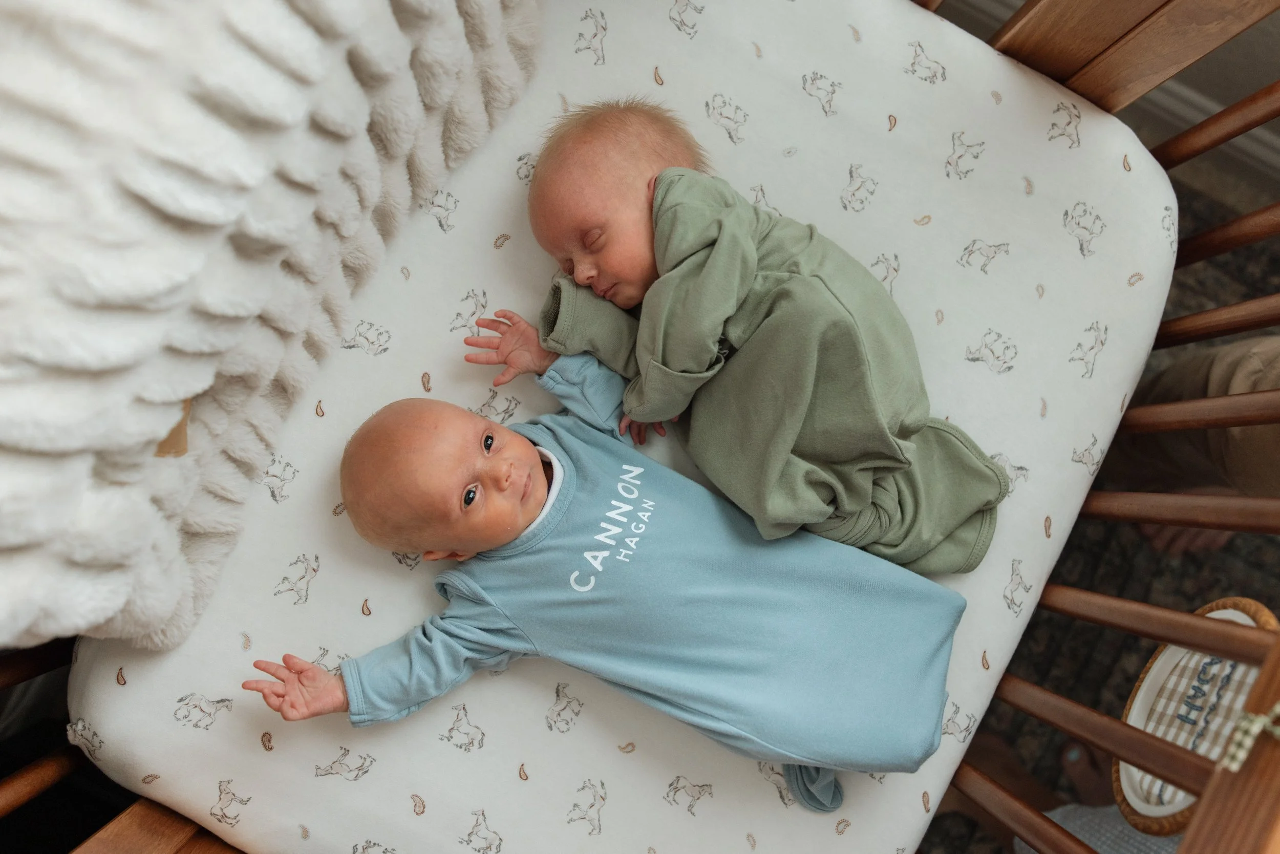 Two young children lying on a bed with a patterned sheet, one is awake and making a peace sign, the other is sleeping. There is a fluffy blanket at the head of the bed. in home newborn photography Austin Texas