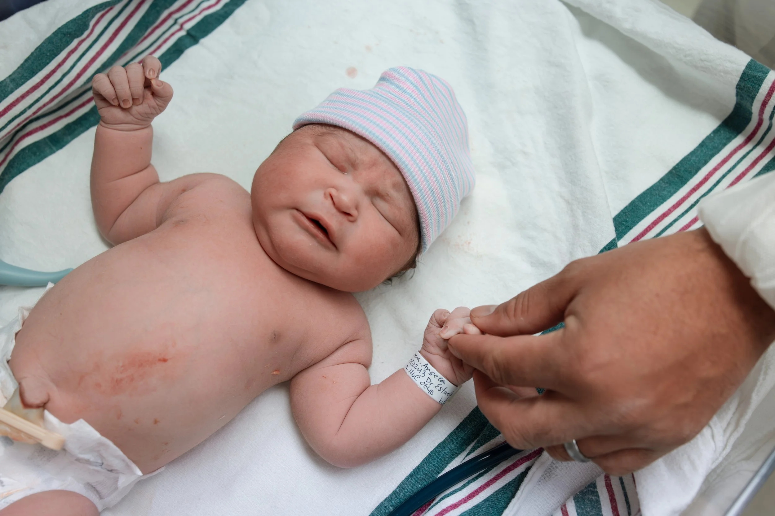 A newborn baby lying on a hospital bed, wearing a pink and blue striped cap, with medical staff holding its hand. The baby has a slightly open mouth and closed eyes. Austin brith photographer 
