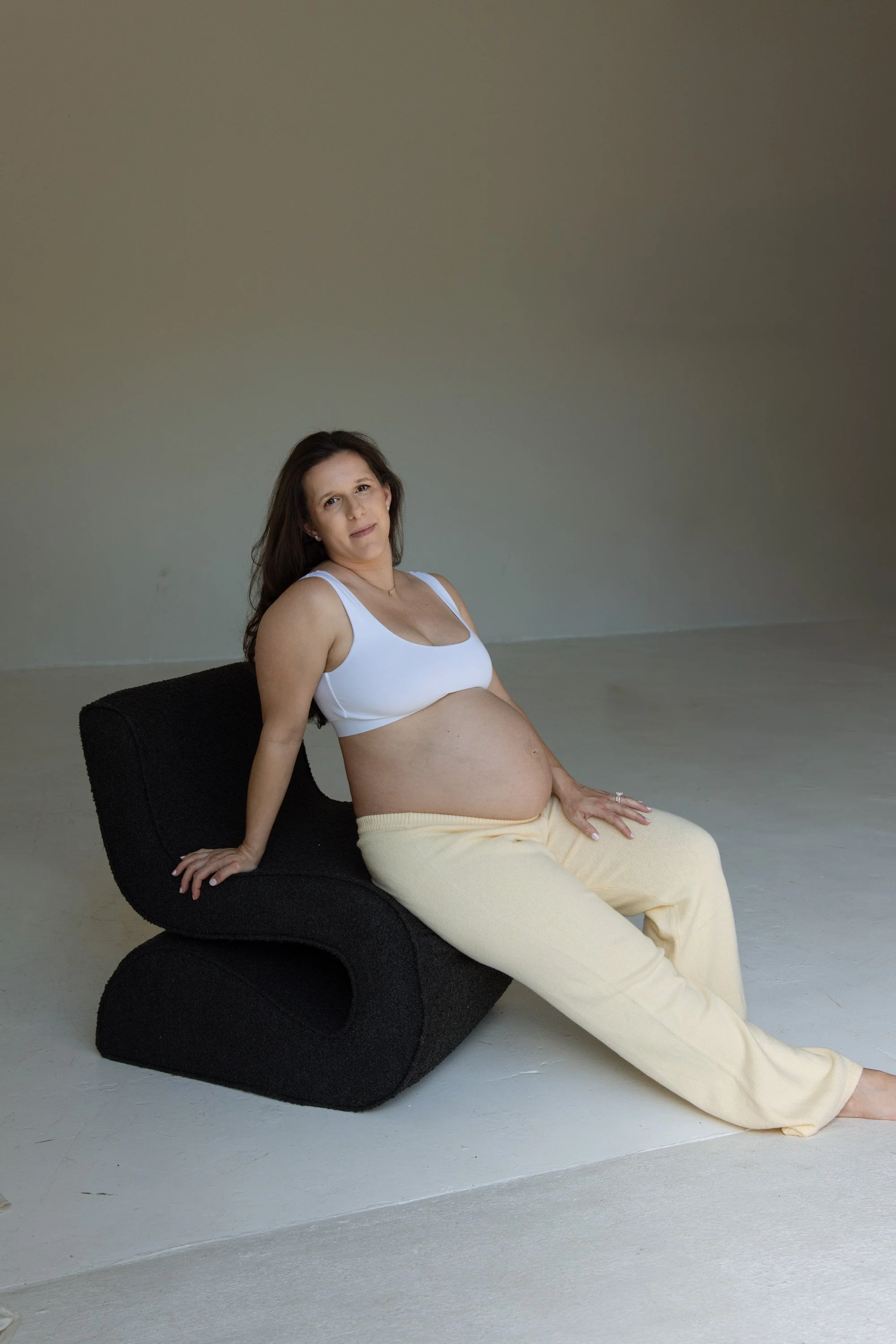 A pregnant woman with long brown hair in a white tank top and yellow pants sits on a black chair against a plain, gray background. She is looking into the camera, her left hand resting on her lap, and her right hand on the chair. editorial studio mat