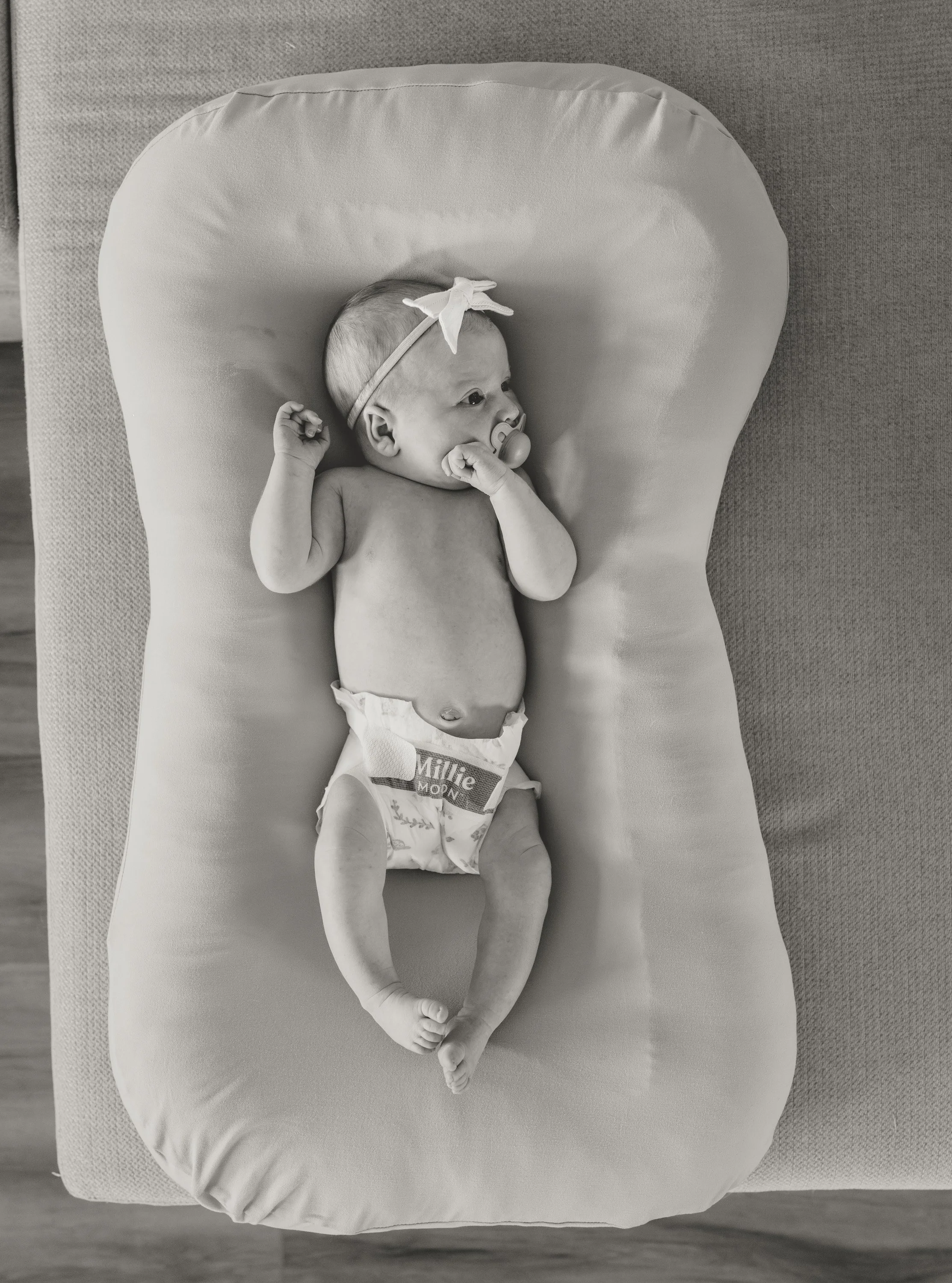 Black and white photo of a baby lying on a cushioned changing pad, with a pacifier in its mouth and a headband with a bow. The baby is wearing a diaper and lying on its back. documentary newborn photography Austin Texas 