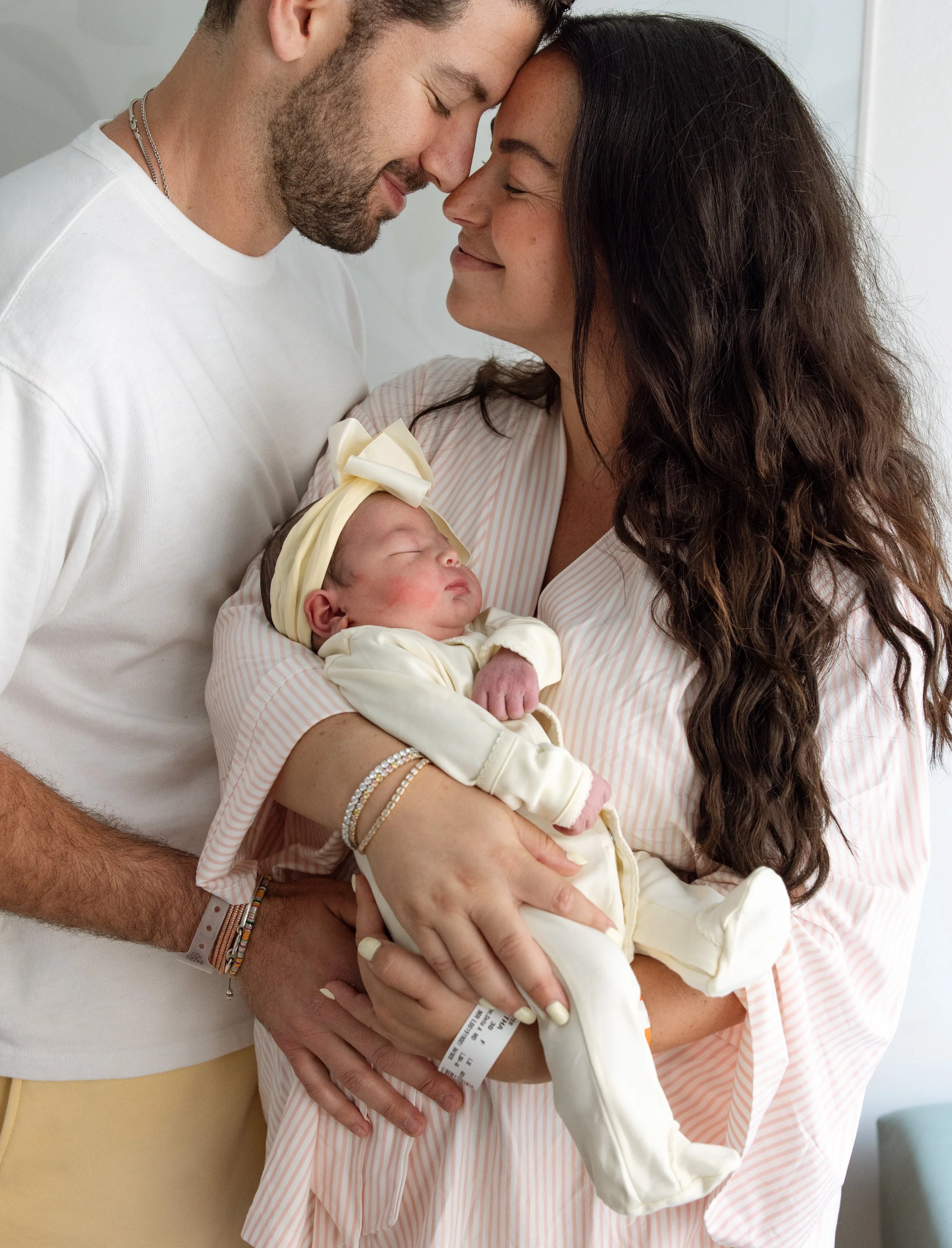 A couple, both with closed eyes and smiling, touch their foreheads together while holding a newborn baby. The woman is wearing a pink and white striped hospital gown, and the man is wearing a white t-shirt and has bracelets on his wrist. The baby is dressed in cream-colored clothing with a bow headband and is being cradled in the woman's arms.