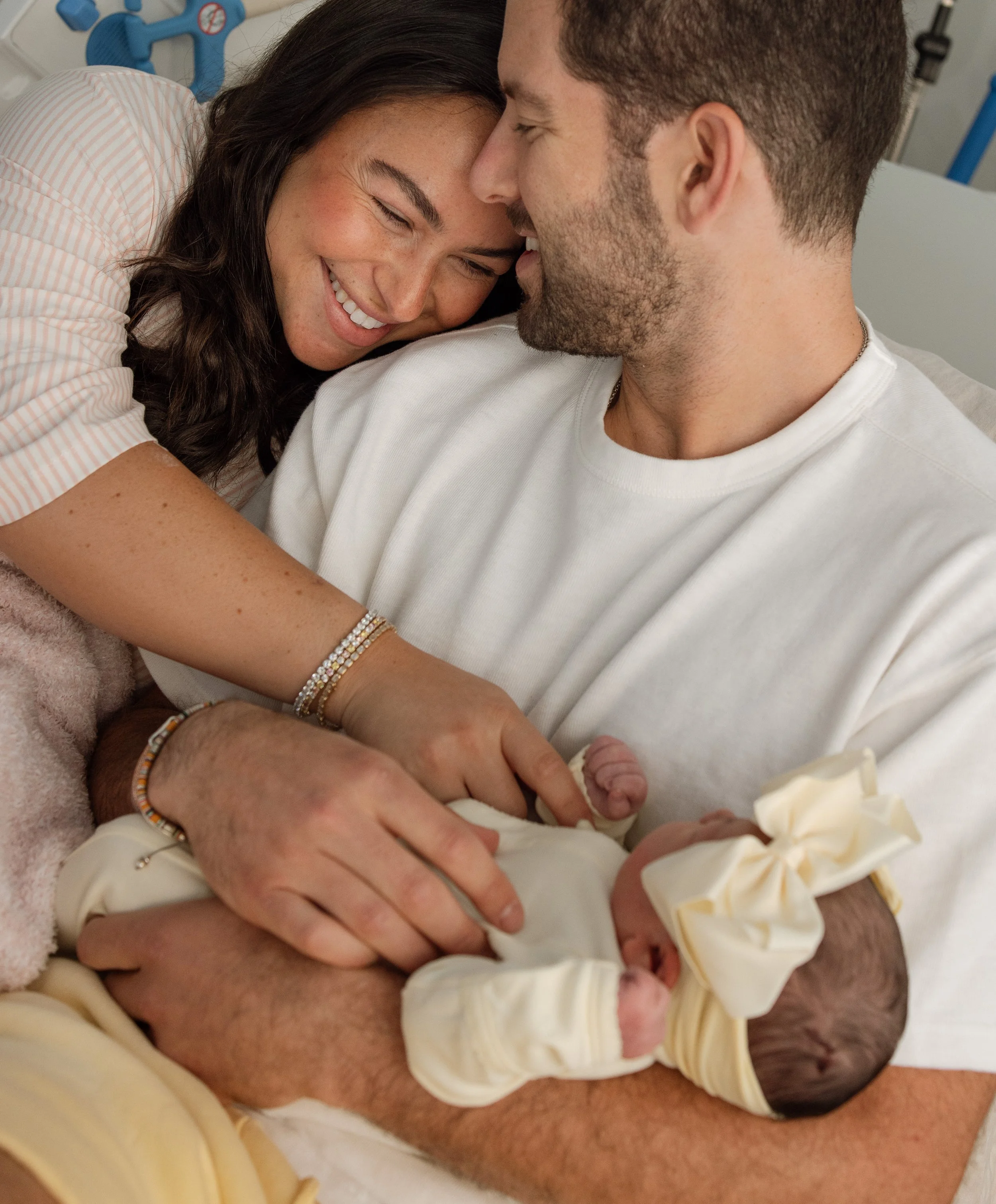 A woman and a man in a hospital bed. The woman appears to be happy and is smiling, leaning in close to the man. The man has a beard and is wearing a white shirt, and they are cuddling or possibly cuddling a newborn baby wrapped in a yellow blanket. There are medical equipment and a blanket visible in the background, indicating they are in a hospital setting.