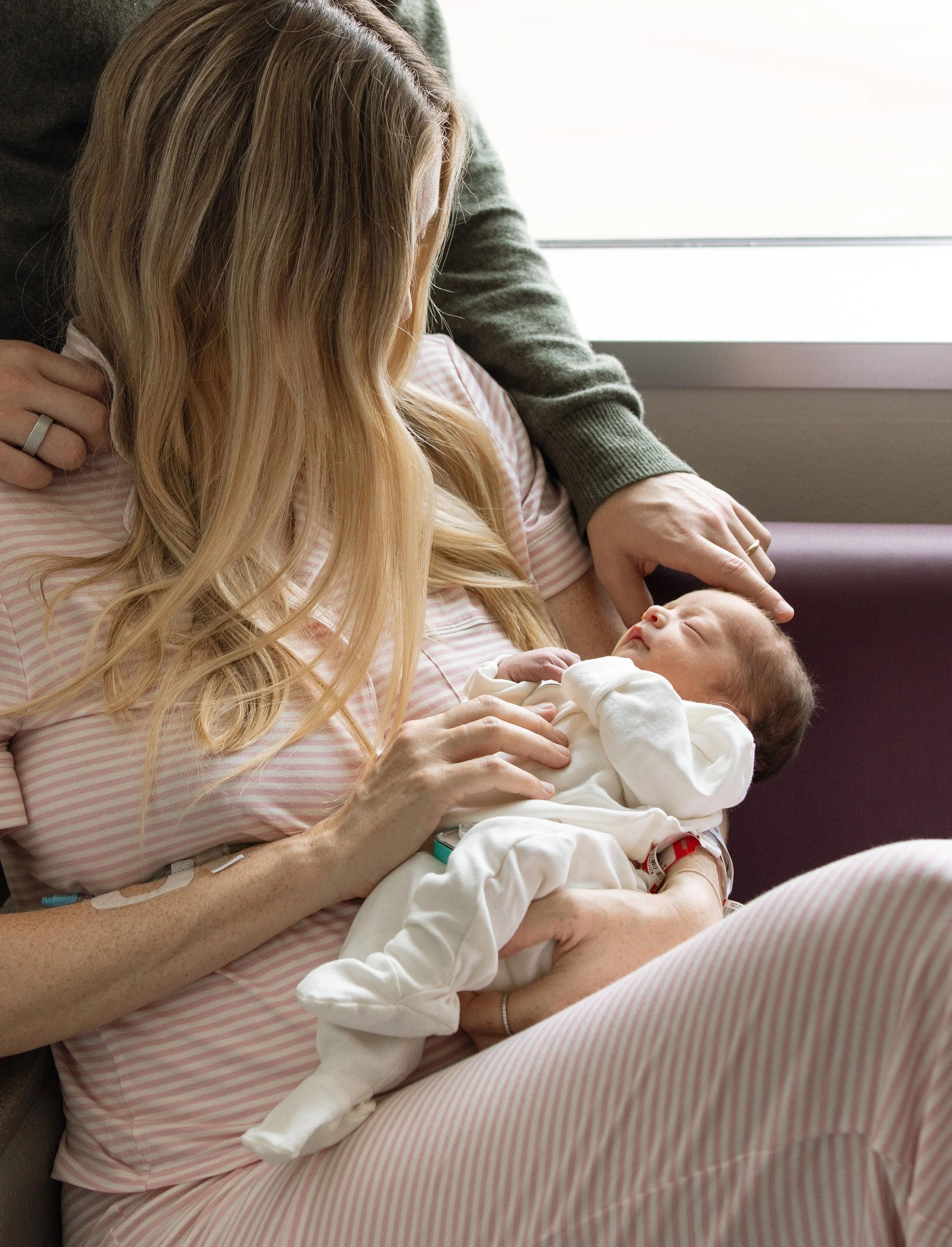 A woman with long blonde hair holding a newborn baby in her arms in a hospital room, with a person standing beside her. The woman is wearing a pink and white striped hospital gown, and the baby is dressed in a white outfit. There is a window with nat