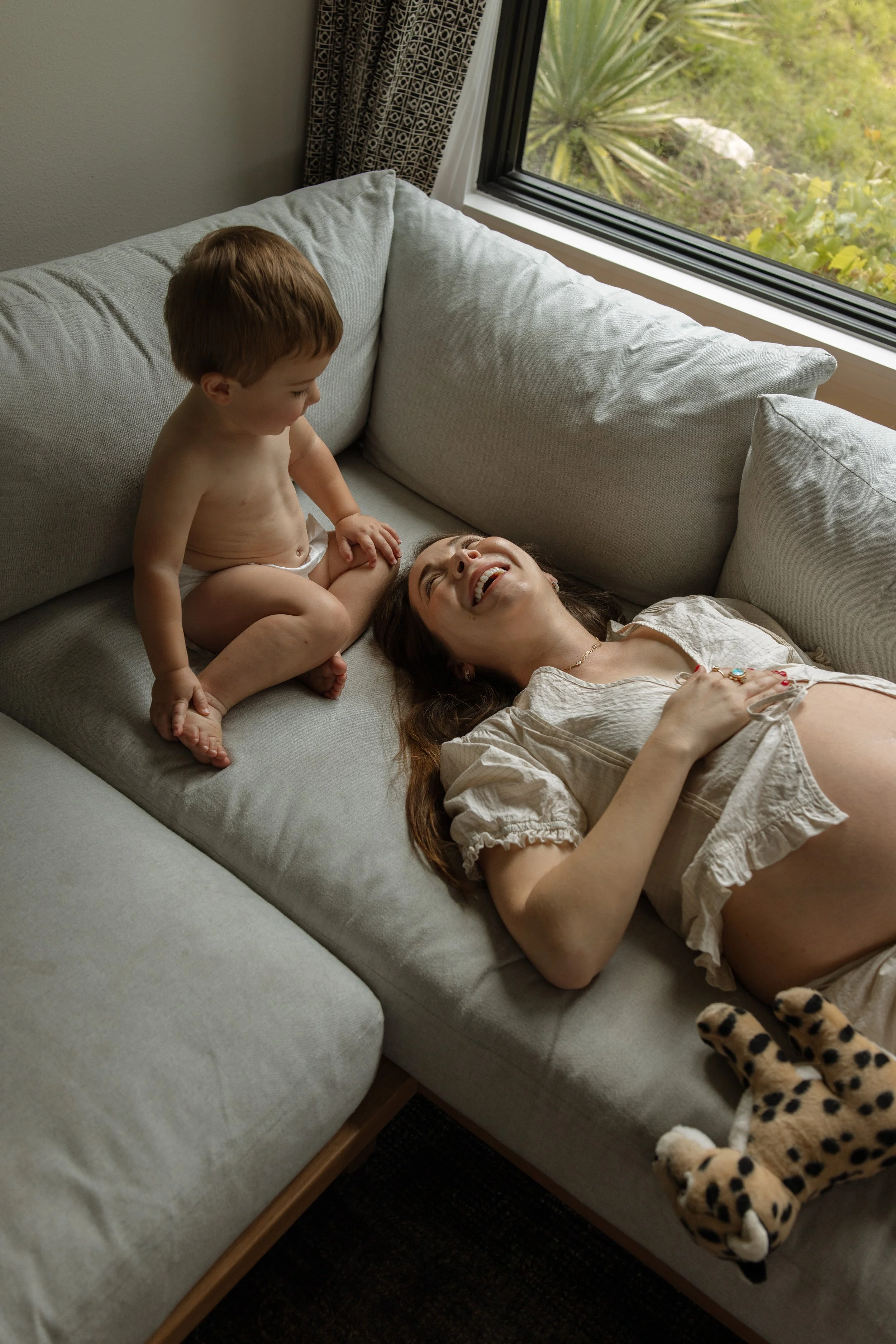 A pregnant mom lying on a light gray sofa, smiling, with a young boy sitting on the armrest beside her. The boy is shirtless, wearing a diaper, and looking at the woman. There is a window in the background showing green foliage, and the woman is wear