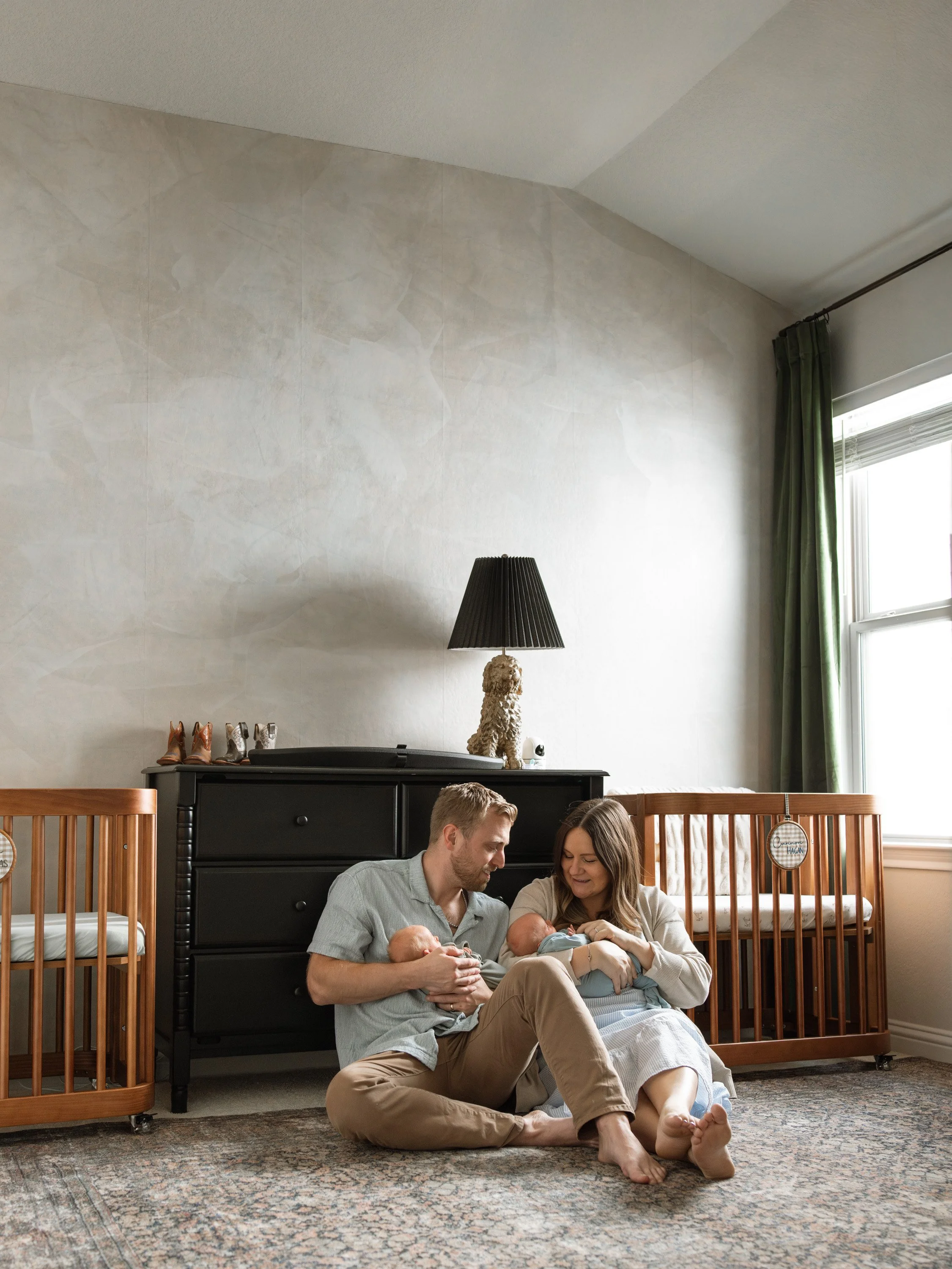 A couple with two babies sitting on a rug in a nursery, smiling at each other and the babies, with a black dresser, a lamp with a dog-shaped base, a wooden crib, and a window with green curtains in the background. newborn photography in Austin Texas