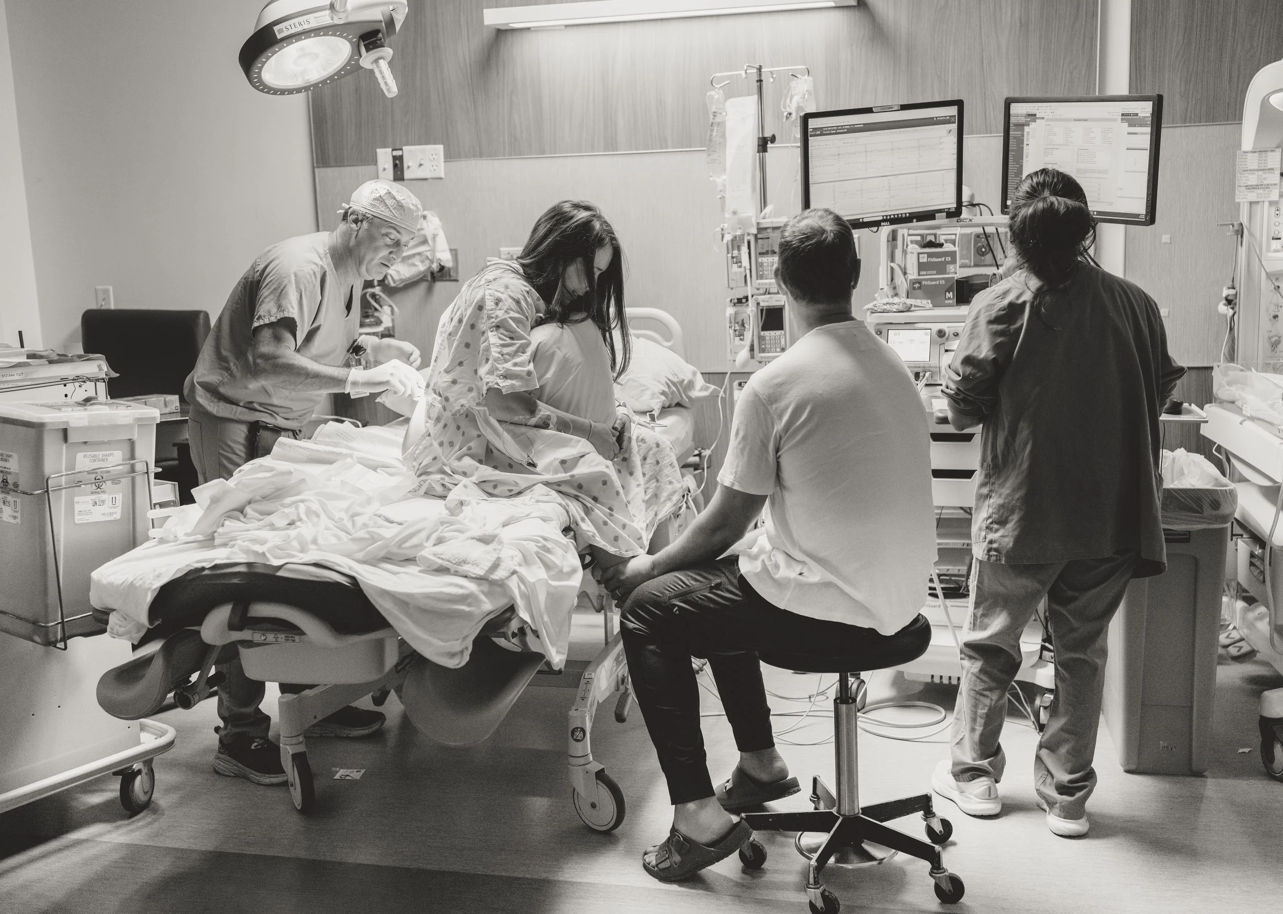 black and white Austin birth photography image shows a pregnant woman in the hospital getting an epidural while her husband, a labor and delivery nurse, and the doctor are all near her.