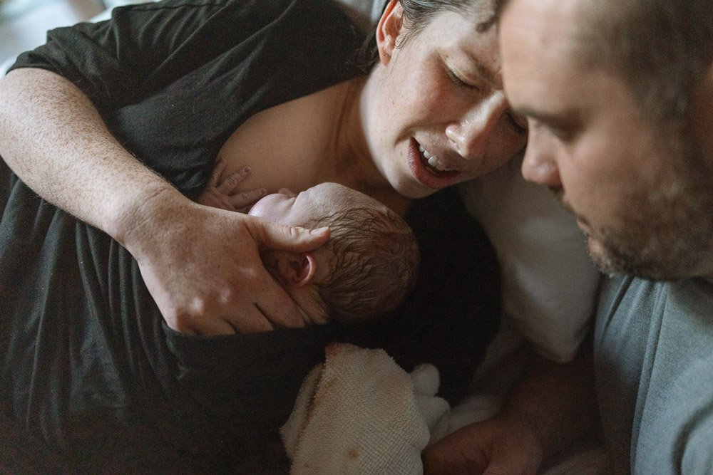 A woman holding a newborn baby close to her chest while lying in bed, with a man leaning in nearby, all appearing to share a moment of affection.