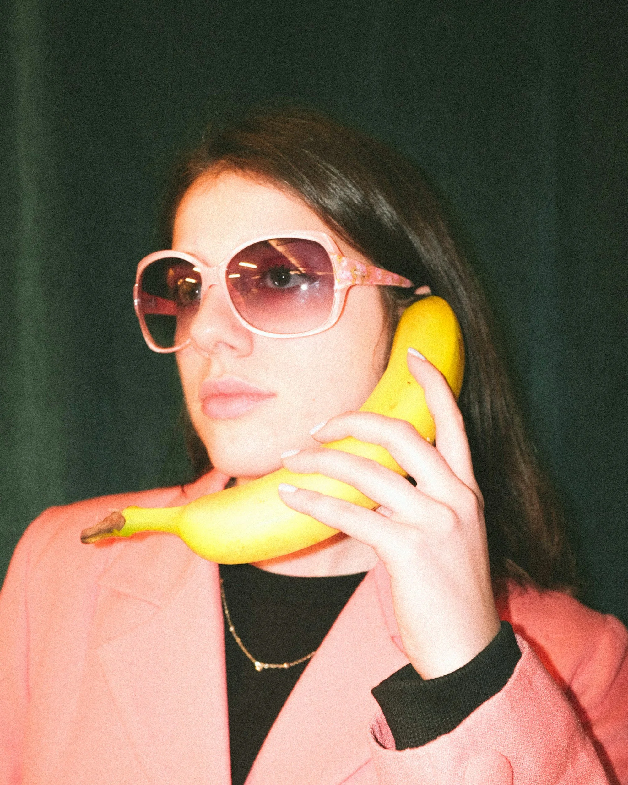 A woman with dark hair wearing pink sunglasses with heart-shaped frames holding a yellow banana to her ear like a phone, against a dark background.