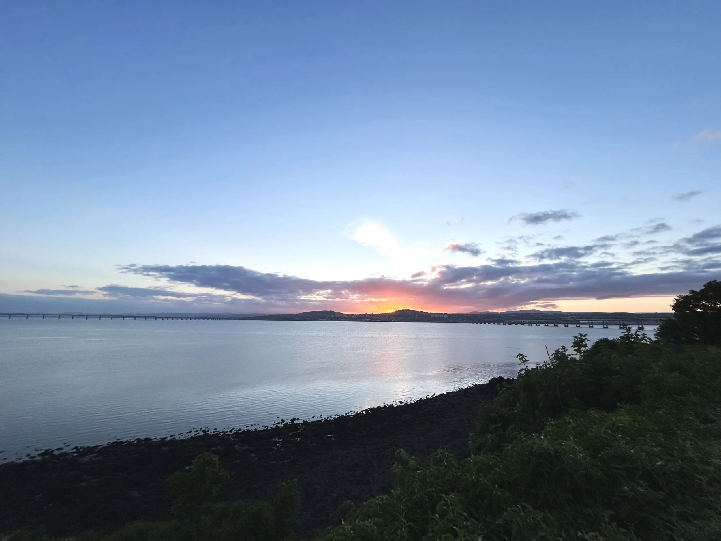 A body of water with a bridge in the distance, a sunset, and clouds in the sky, with trees and rocks along the shoreline. tay firth taken from Newport on tay