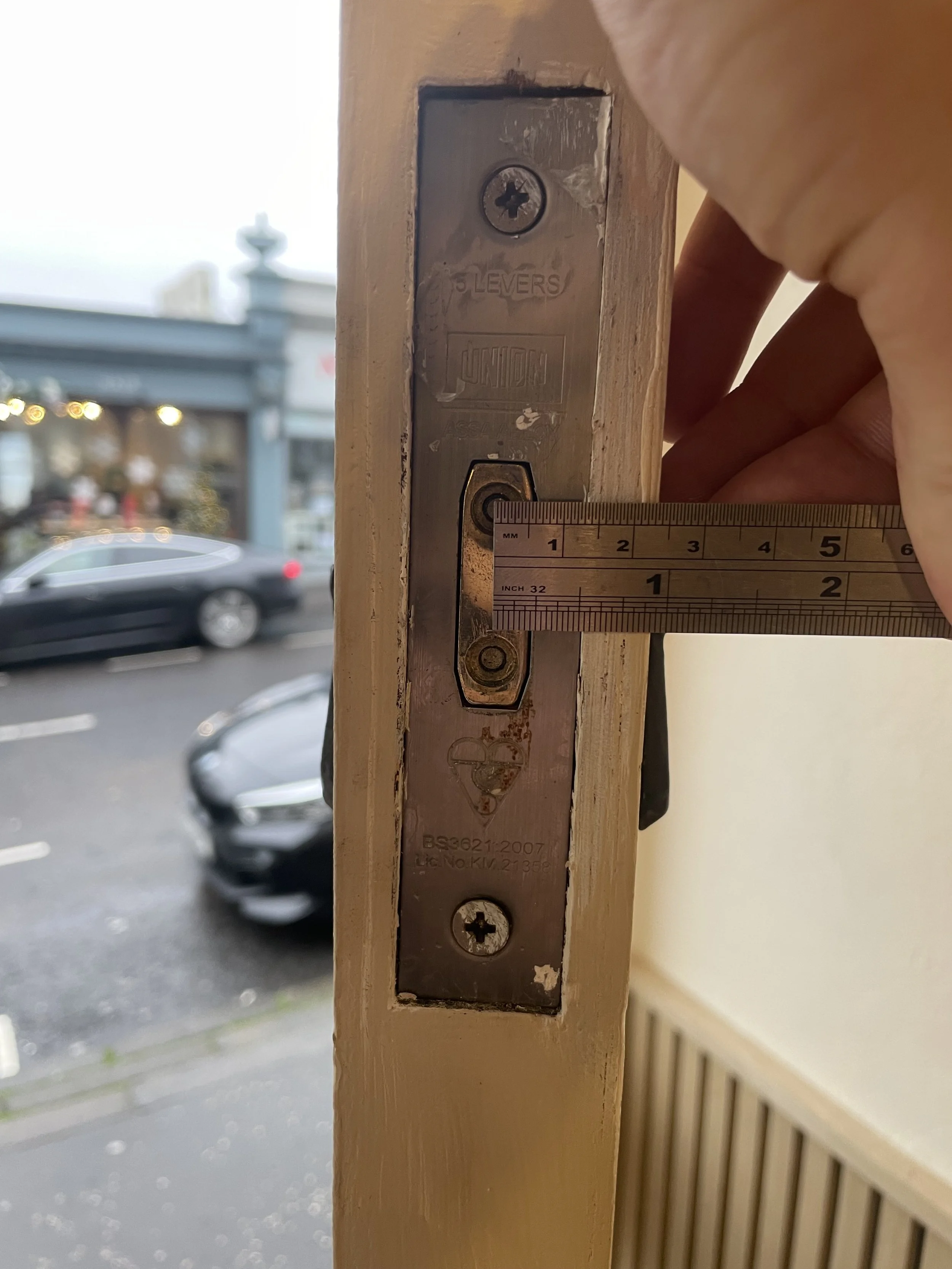 Close-up of a metal door latch plate with a screw in a wooden door frame, with a blurred parking lot and cars visible outside.