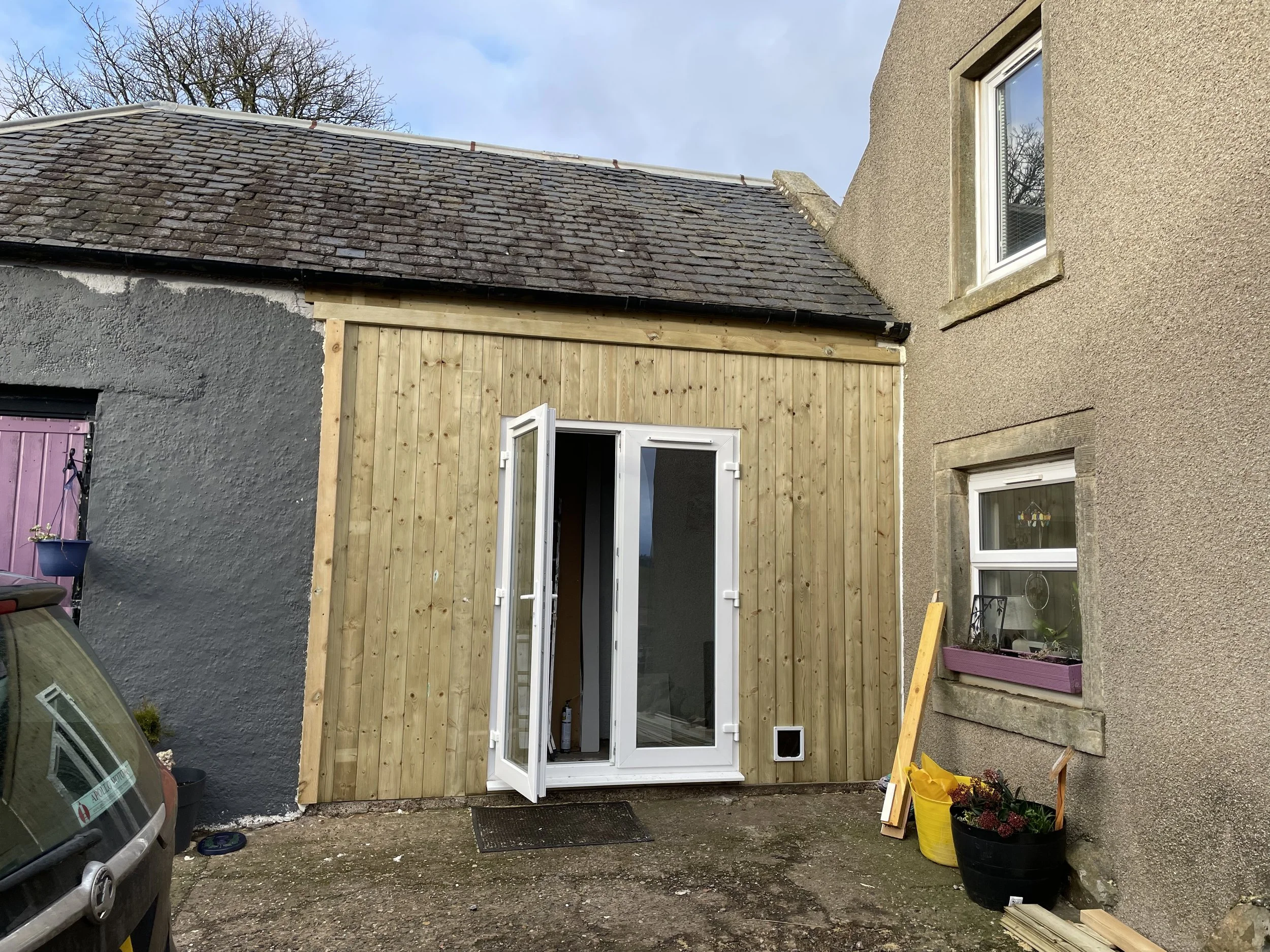 Backyard construction scene with a partially built wooden extension on a house, featuring an open door and a window, with construction materials and flower pots on the ground. built by Gallier Locksmithing. patio door and wooden partition.
