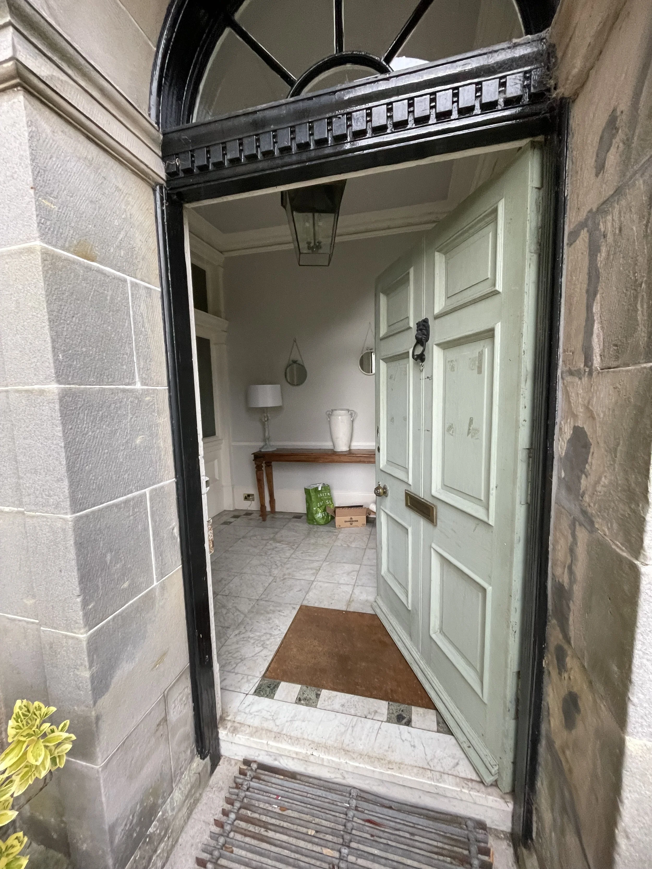 View of a building entryway with a green door slightly ajar, a doormat, and a small array of pipes on the ground. The image is taken from outside, looking in through an open door, with stone and brick walls surrounding the entrance and a mirror and table visible inside.