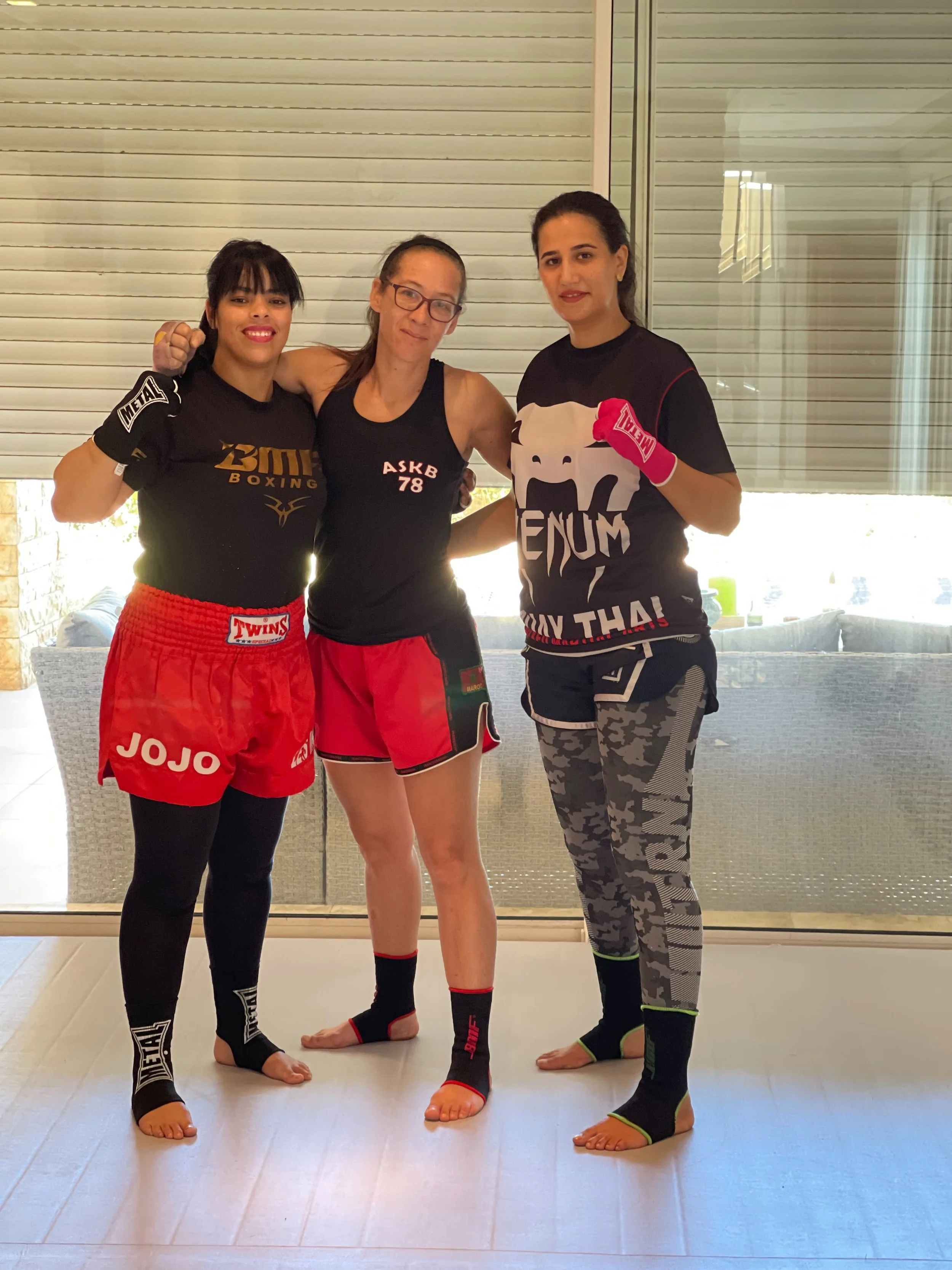 Trois femmes posant en tenue de boxe thaï sur un tapis d’entraînement, avec gants et shorts de combat.