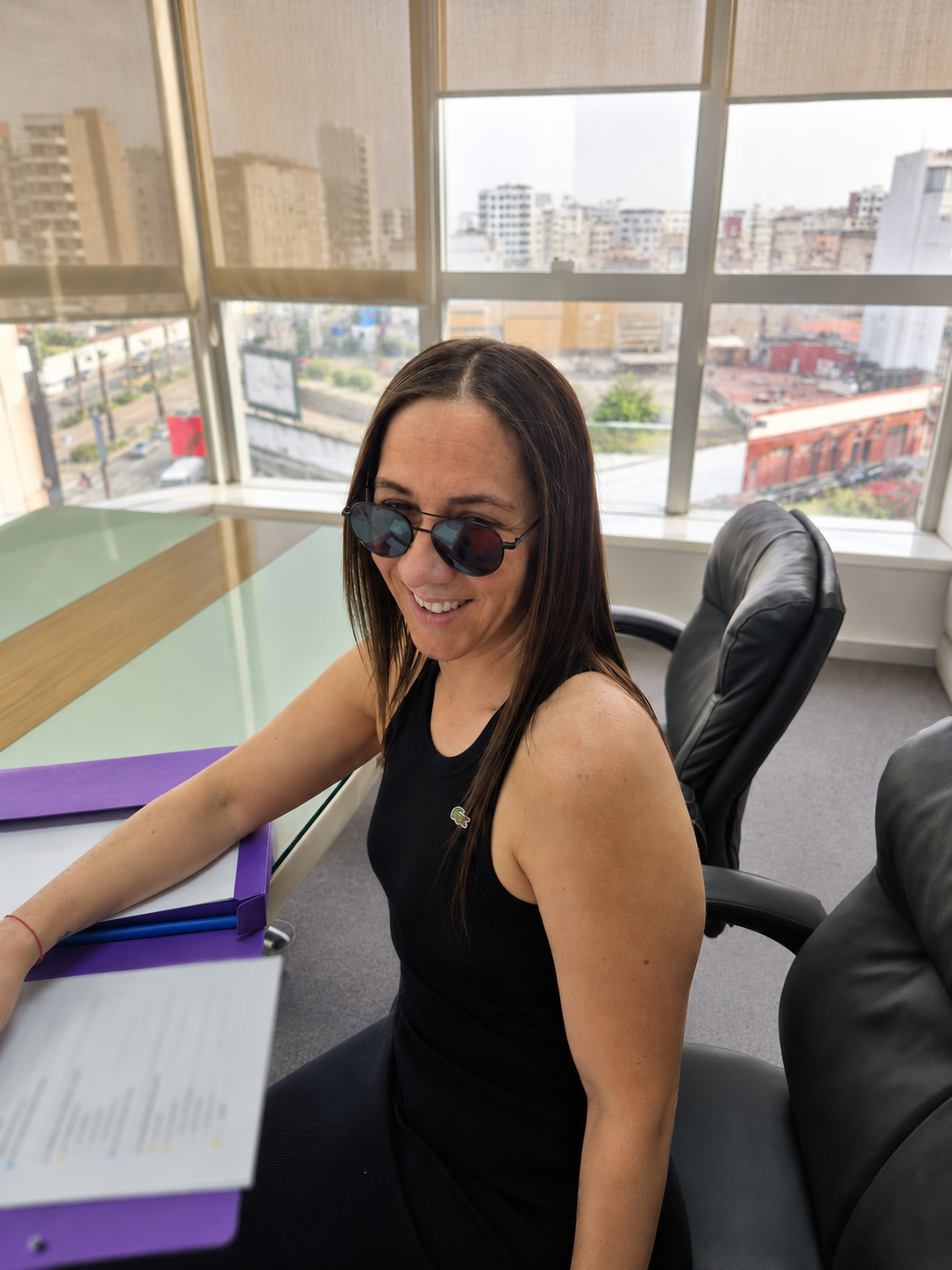 Femme assise à une table de bureau, portant des lunettes de soleil, souriant, avec des fenêtres donnant sur une ville en arrière-plan.