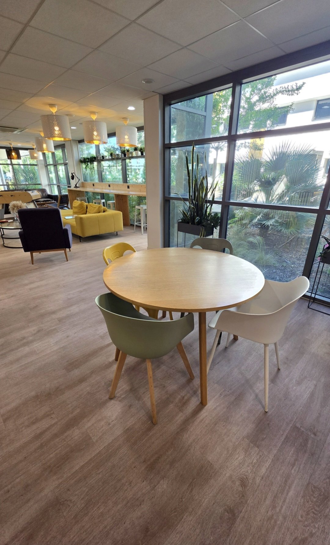 A round wooden table surrounded by five modern chairs in a bright, spacious room with large windows, outdoor greenery visible outside, and a living area with sofas and plants in the background.