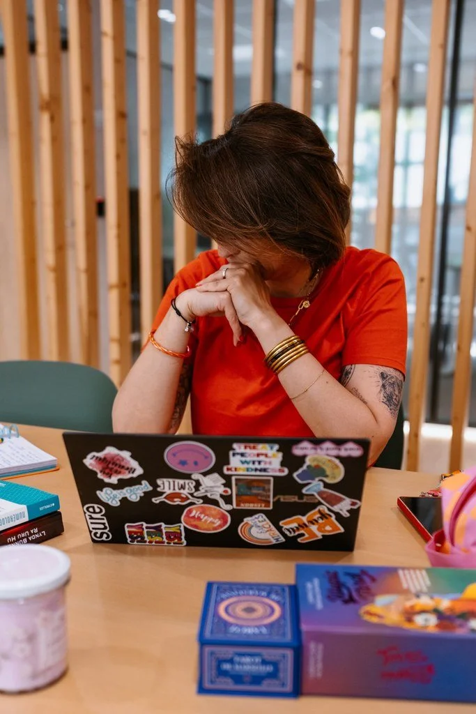 A woman wearing an orange shirt is sitting at a desk with her head bowed and hands clasped, in front of a decorated laptop. The workspace has various items including a tablet, a box of cards, and a container of pink ice cream. The background features wooden wall panels and large windows.