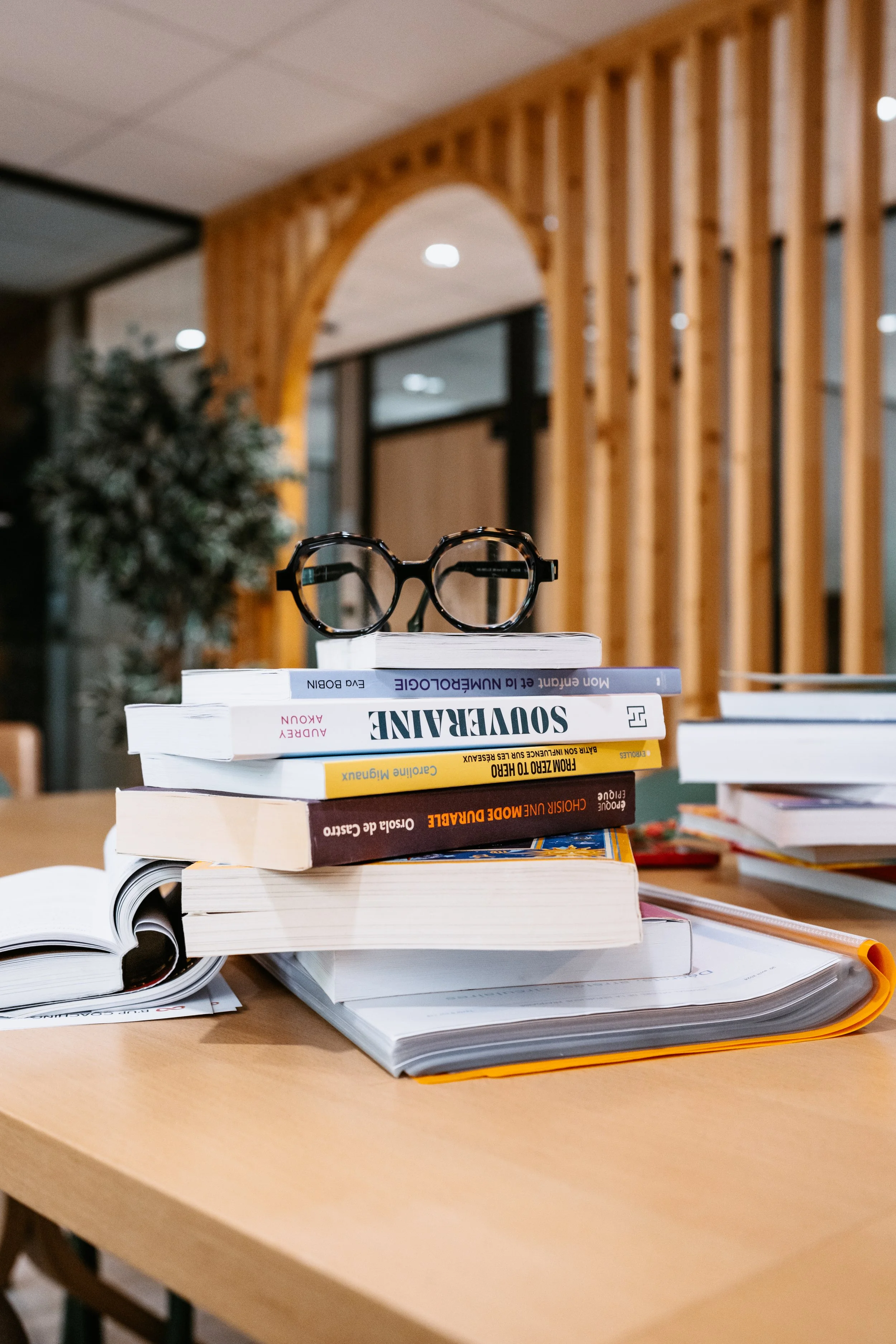 A stack of books with a pair of black eyeglasses on top, placed on an open notebook on a wooden table in a modern interior setting.