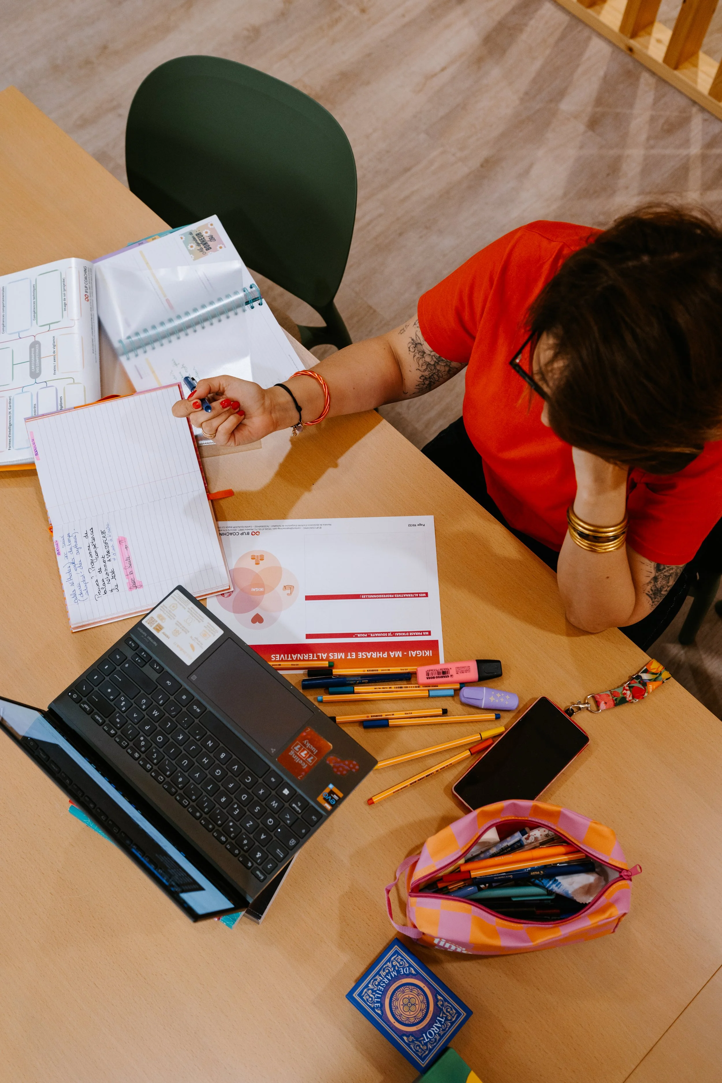 Overhead view of a cluttered desk with notebooks, pens, a laptop, a smartphone, and a small pink backpack filled with pens and markers. A person wearing a red shirt, glasses, and bracelets is sitting at the desk, writing in a notebook.