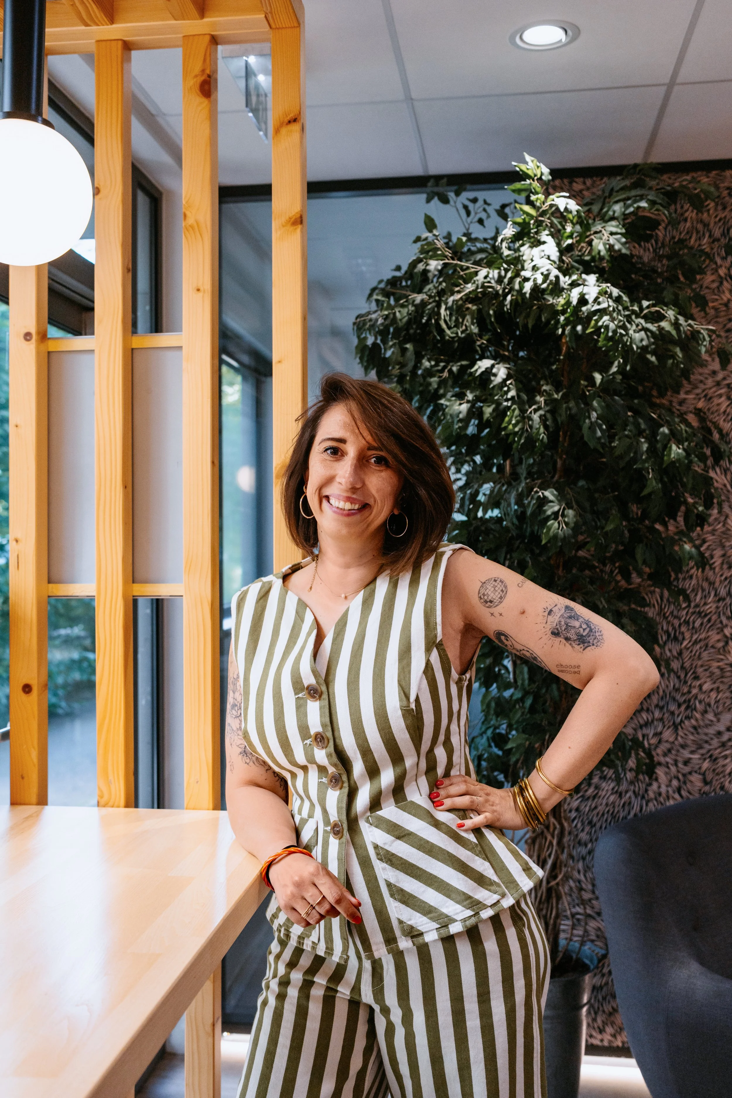 A woman with tattoos on her arms and shoulder, wearing a sleeveless green and white striped button-up shirt and matching pants, smiles at the camera in a modern, well-lit interior with wooden and green decor.