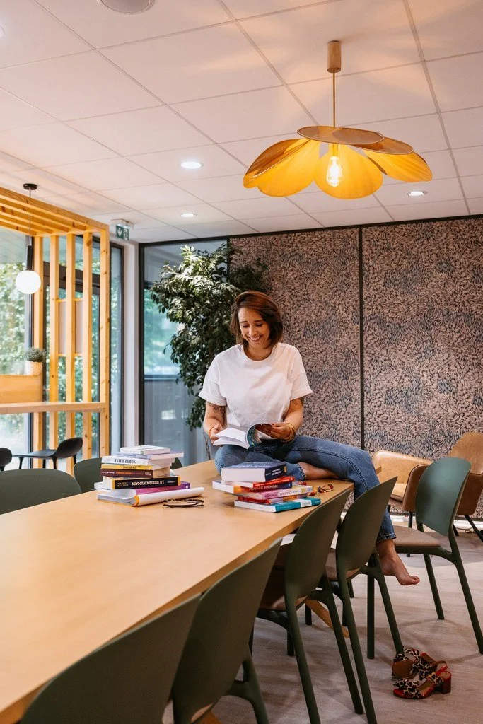 Woman sitting on a conference table filled with books, reading and smiling in a modern, well-lit room with wooden accents and patterned wallpaper.