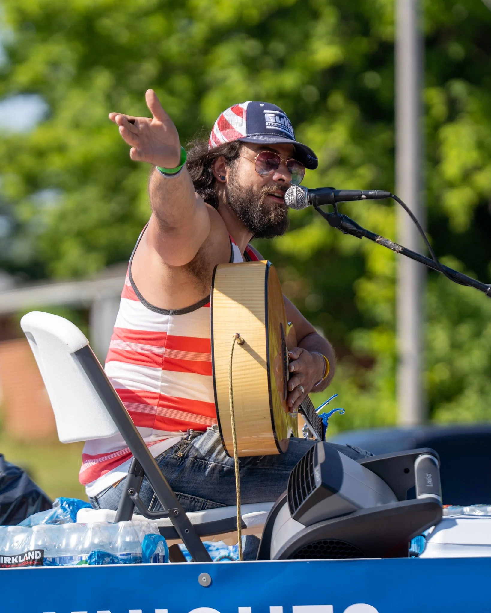 A man with long hair and a beard playing acoustic guitar at an outdoor event, wearing sunglasses, a hat with a checkered pattern, and a sleeveless striped shirt. He is singing into a microphone.