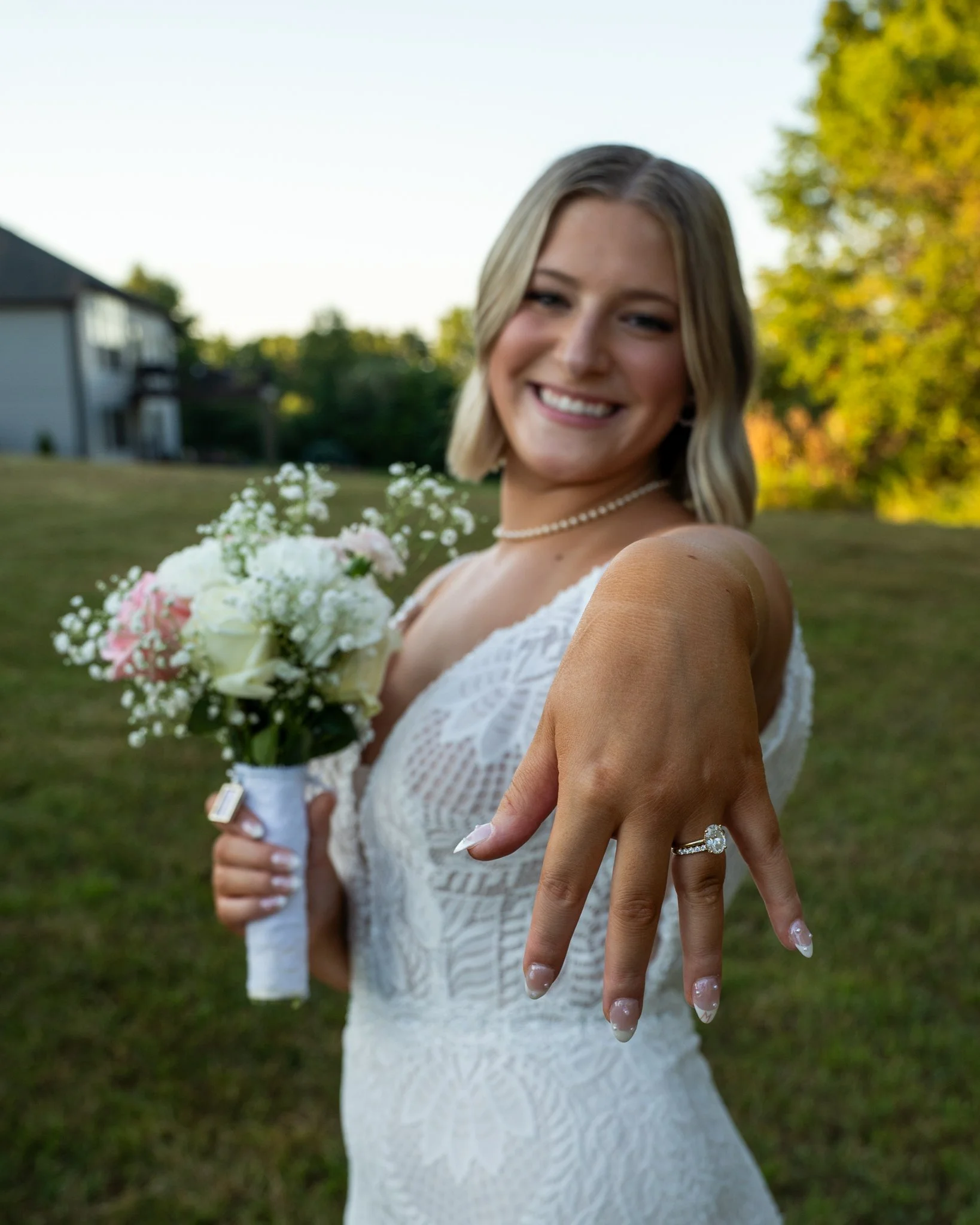 Smiling woman in a white lace wedding dress holding a bouquet of flowers in an outdoor setting with trees and a house in the background.