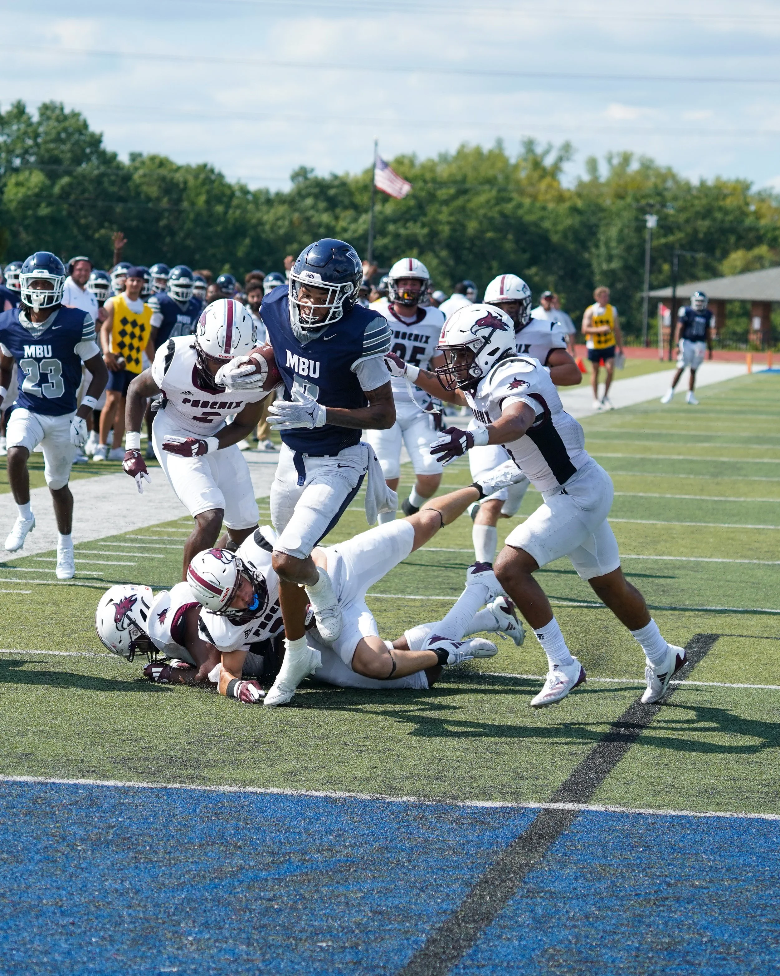 Football game with players in white and navy jerseys on a grass field, some players on the ground and others running, with a background of trees and an American flag.
