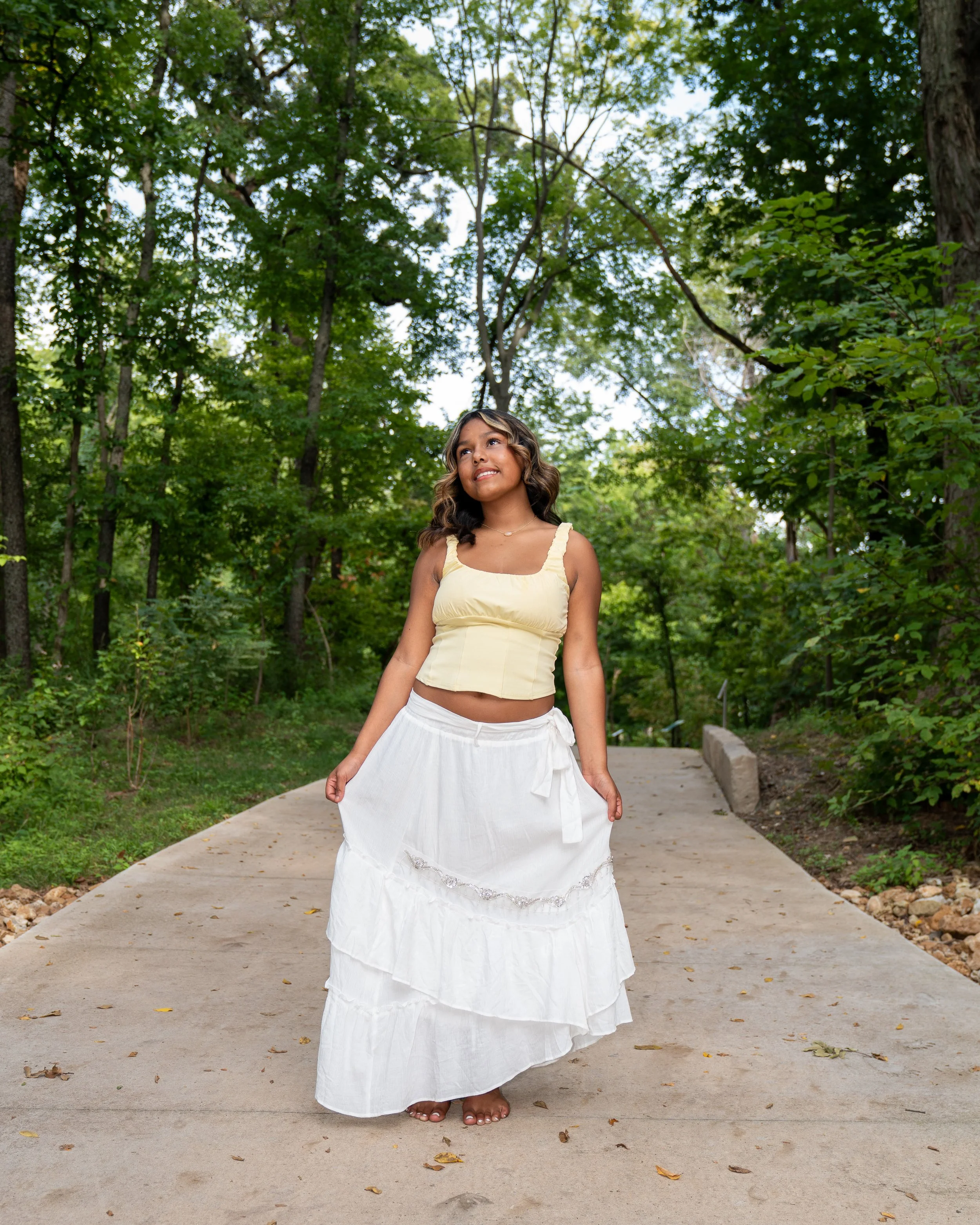 A woman standing barefoot on a concrete pathway in a wooded area, wearing a cream-colored crop top and a long, white flowing skirt, looking up and to her right with a smile.