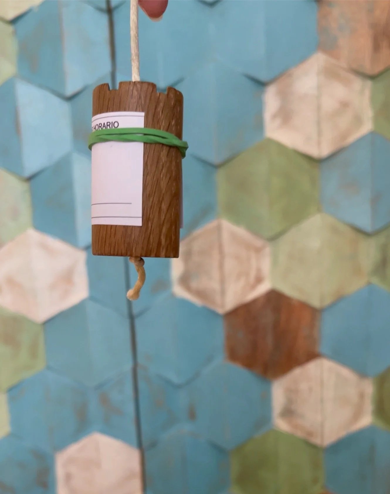 Pendulo Ebraico è un cilindro fatto in legno con etichetta e elastico verde, appeso a un muro con piastrelle colorate a forma di losanga