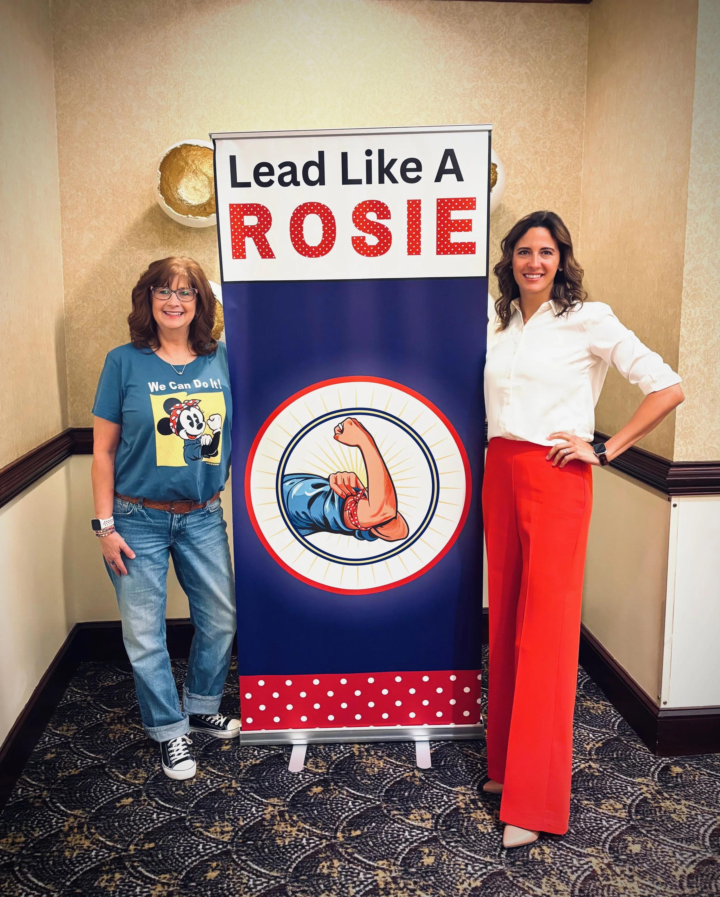 Two women standing on either side of a poster with the text "Lead Like A Rosie," depicting Rosie the Riveter with a flexed arm. The woman on the left is wearing glasses, a blue T-shirt with Minnie Mouse on it, jeans, and Vans sneakers. The woman on the right is wearing a white blouse and red professional pants, smiling with her hand on her hip, and wearing a watch.