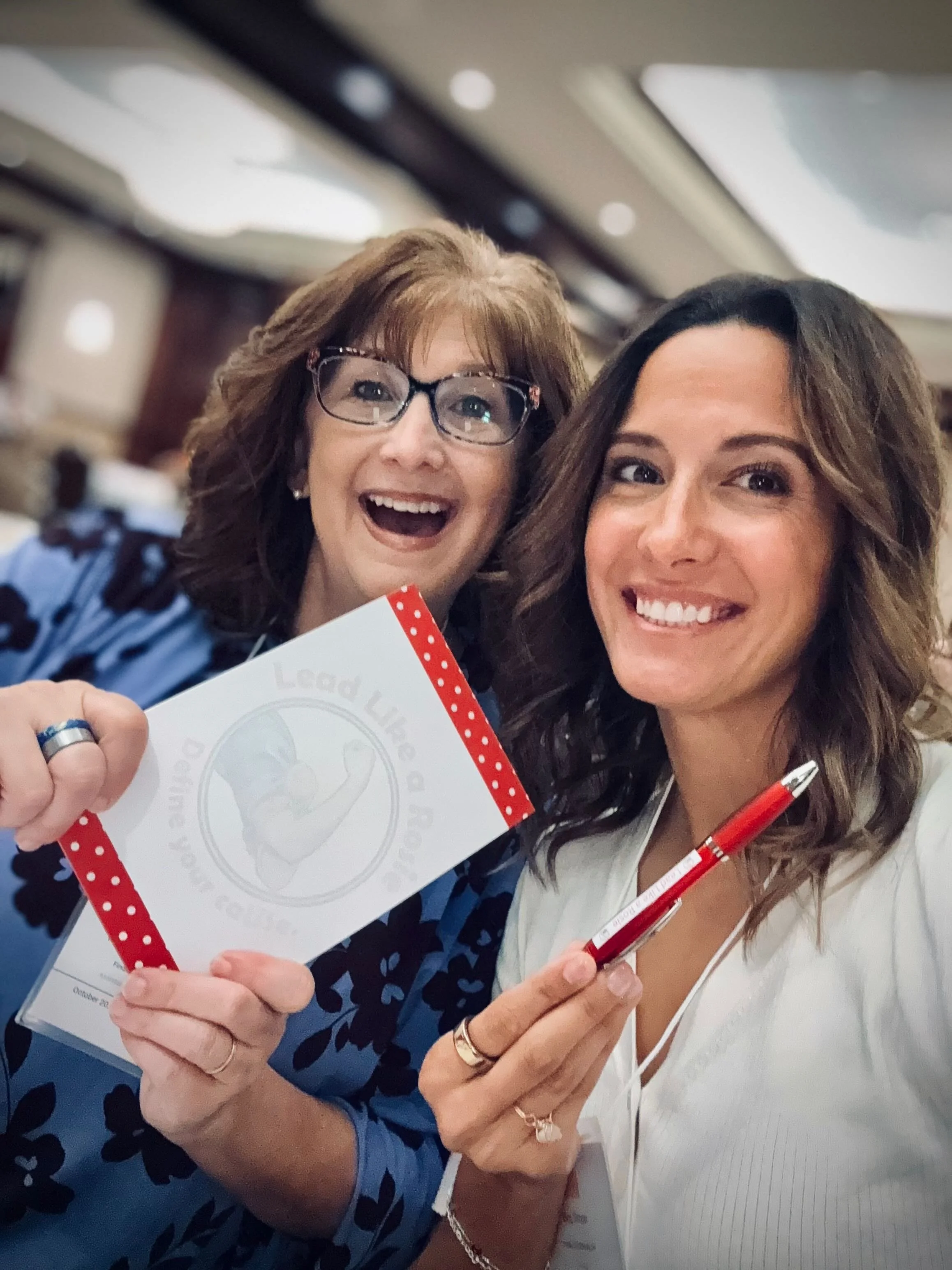 Two women smiling for a selfie, one is holding a certificate and a red pen, indoors at an event.