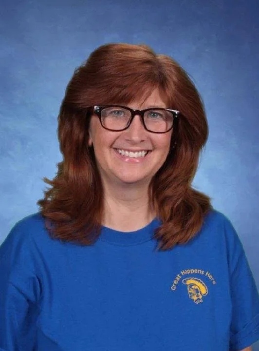 A woman with red hair, glasses, and a blue shirt smiling, standing against a blue background.