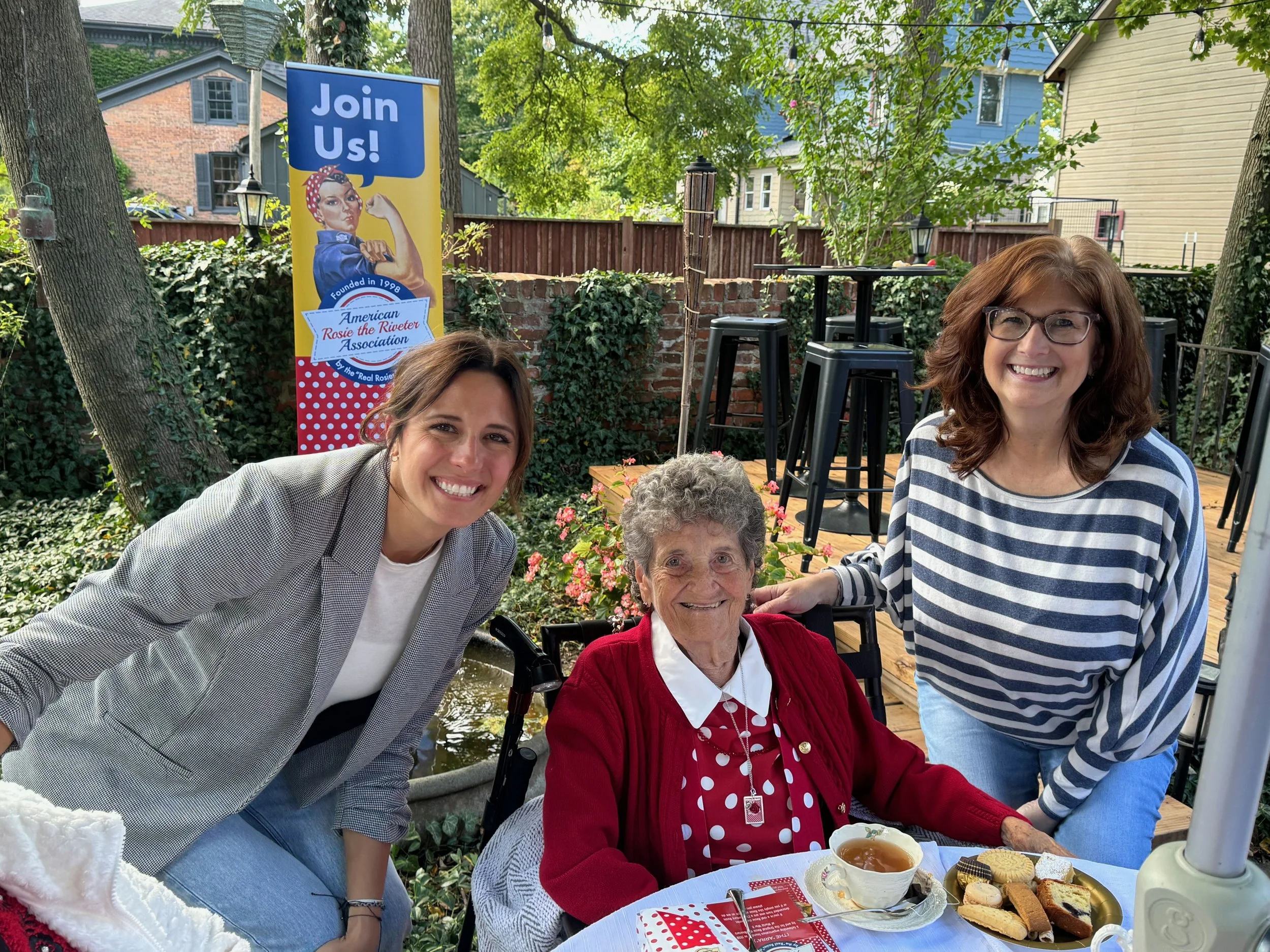 Four women and an elderly woman in a red cardigan sitting at a table outdoors during a garden party with a sign for the American Rosie the Riveter Association. The table has tea, cookies, and desserts.