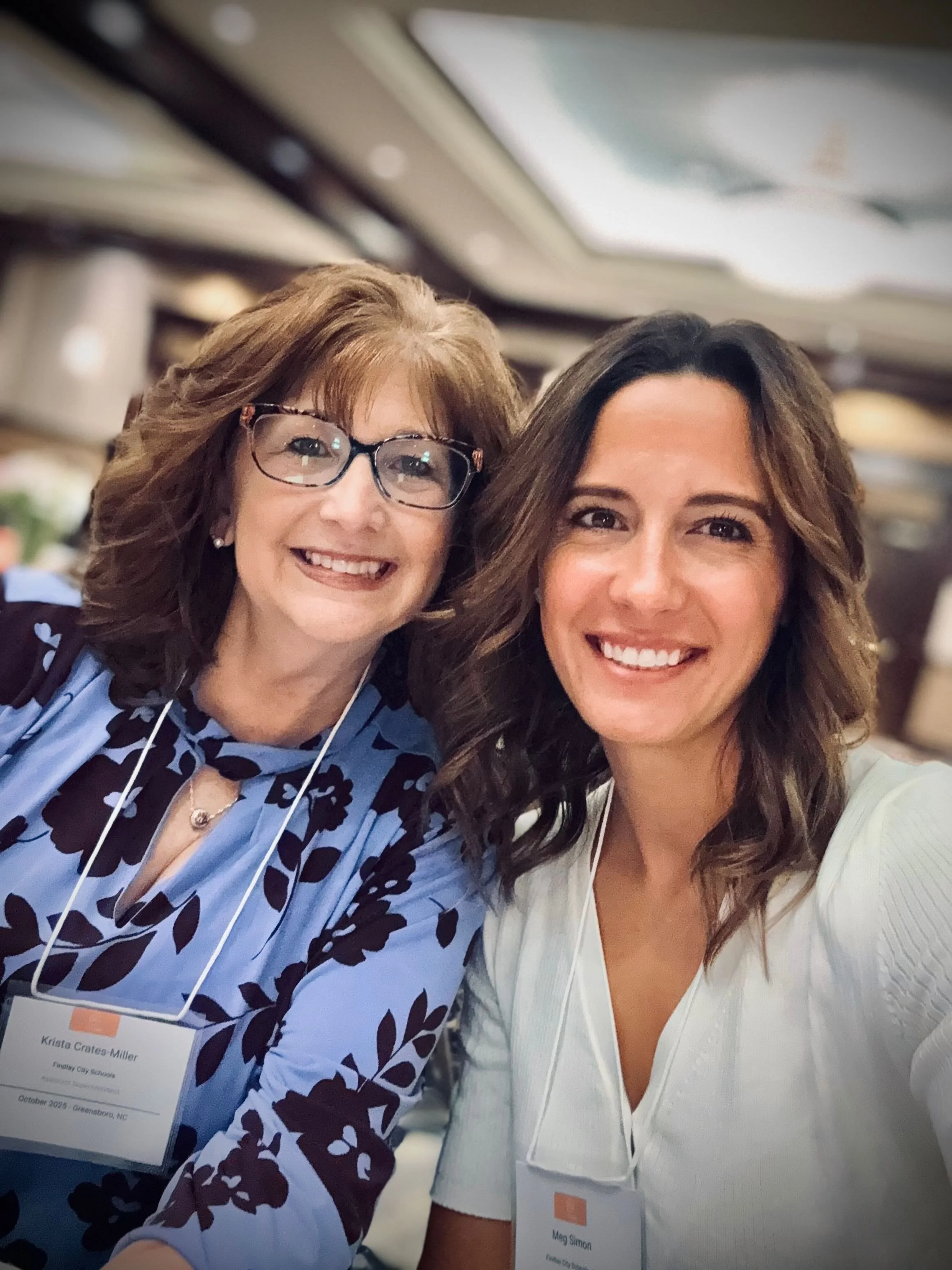Two women smiling for a selfie at an indoor event, each wearing name tags around their necks.